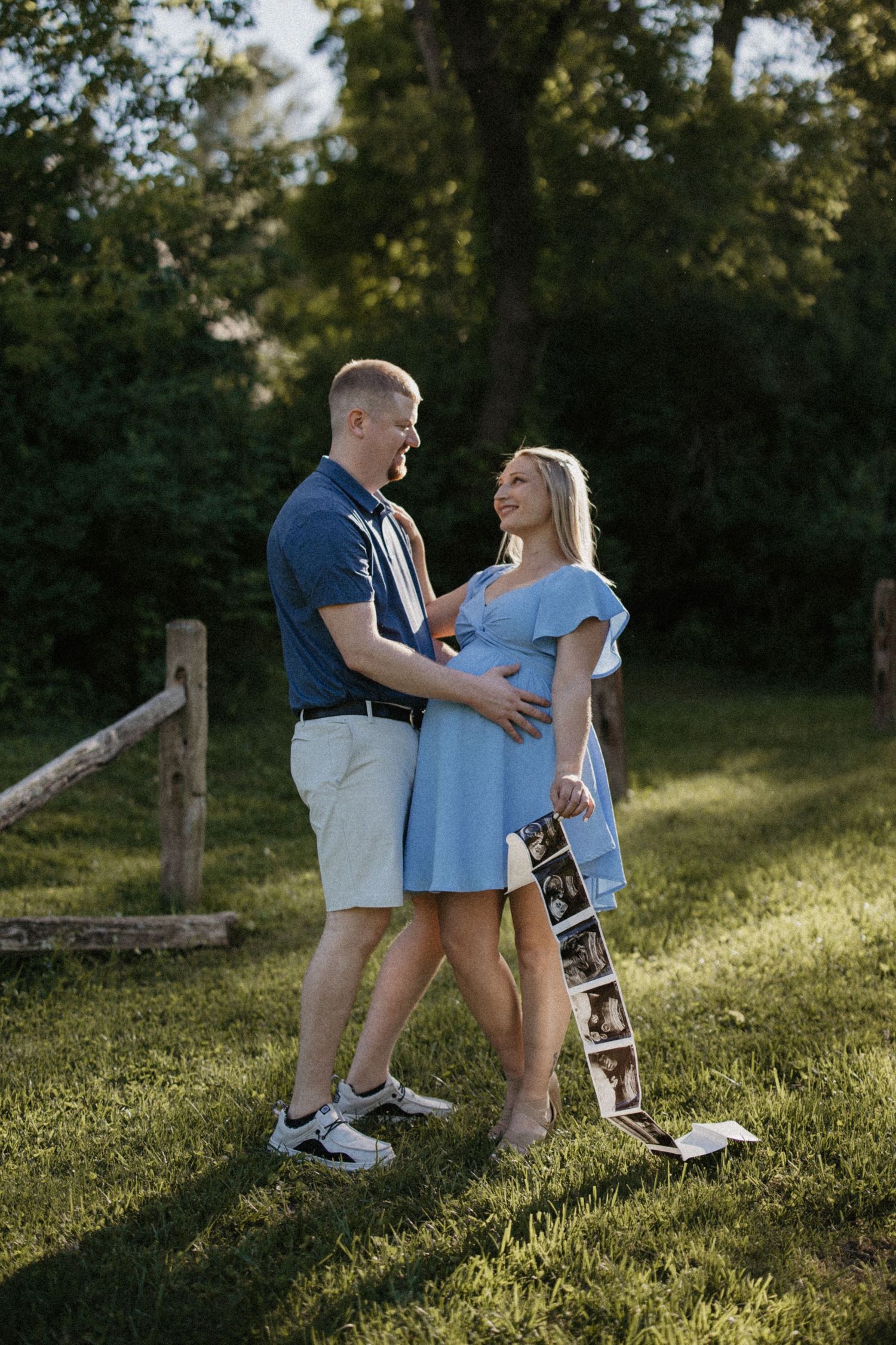 Couple sharing a joyful moment outdoors, woman in blue dress holding ultrasound photos, man touching her baby bump.