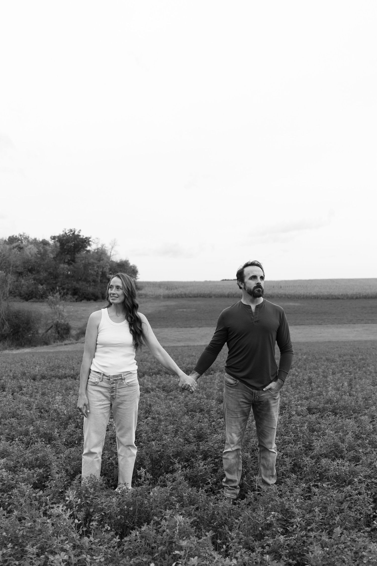 Black and white photo of a couple holding hands in a field, smiling, with trees and sky in the background.
