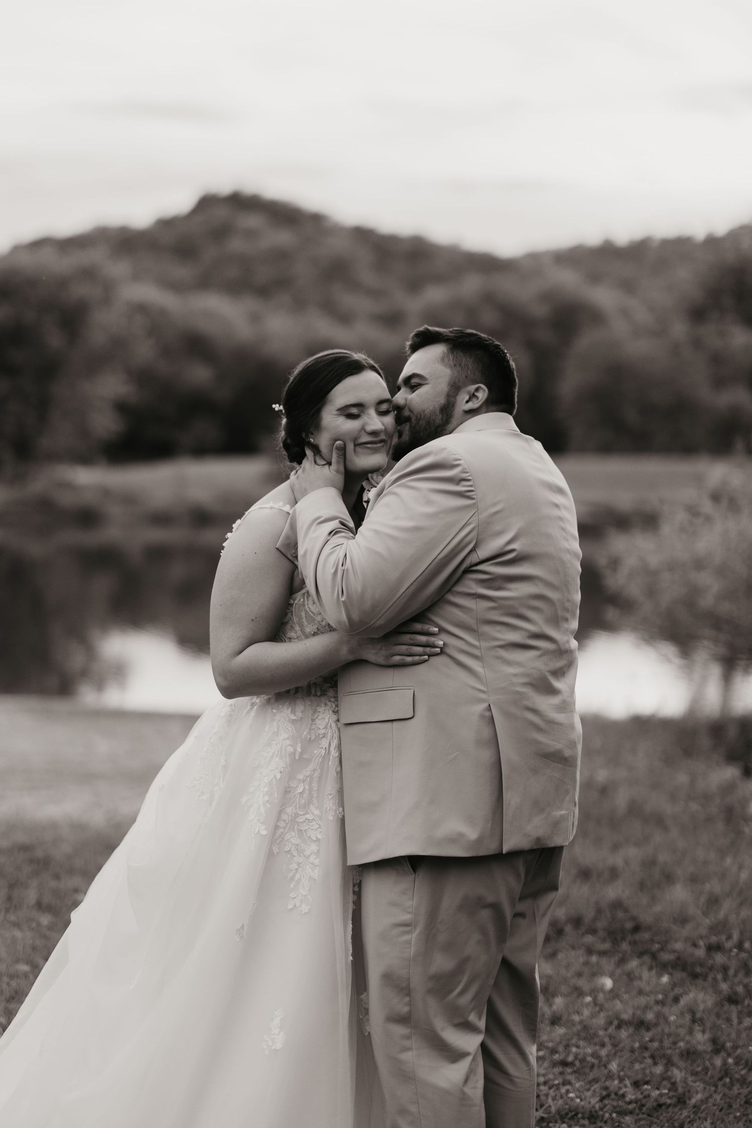Black and white photo of a groom kissing his bride on the cheek outdoors near a pond with rolling hills behind them.