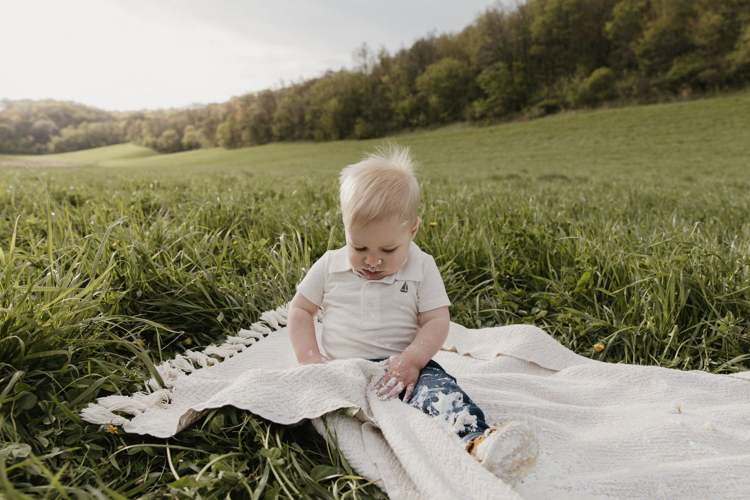 A baby sits on a white blanket in a lush green field, wearing a white onesie and floral pants.