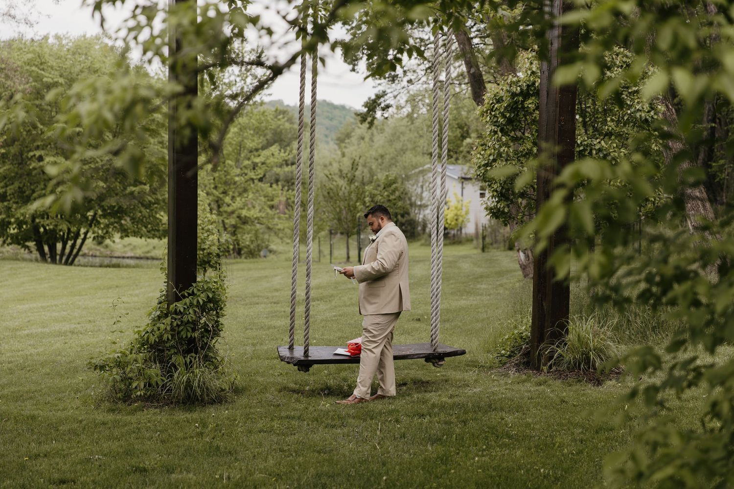 Man in beige suit standing by a wooden swing set in a lush green garden surrounded by trees.
