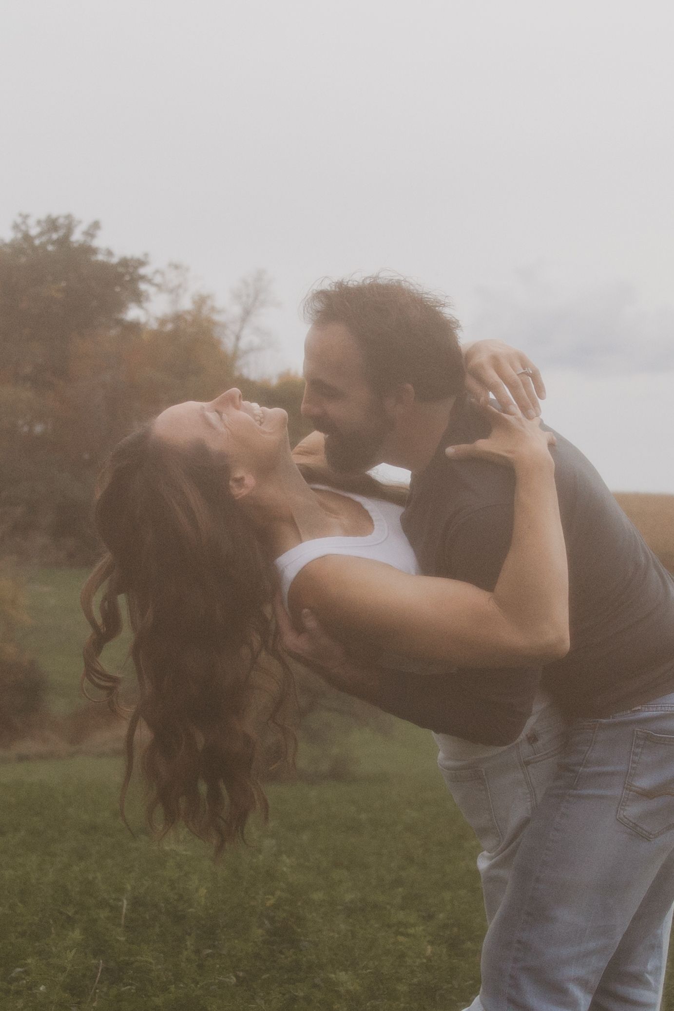 A couple shares a romantic moment outdoors on a misty day, the woman laughing as the man leans in to kiss her.
