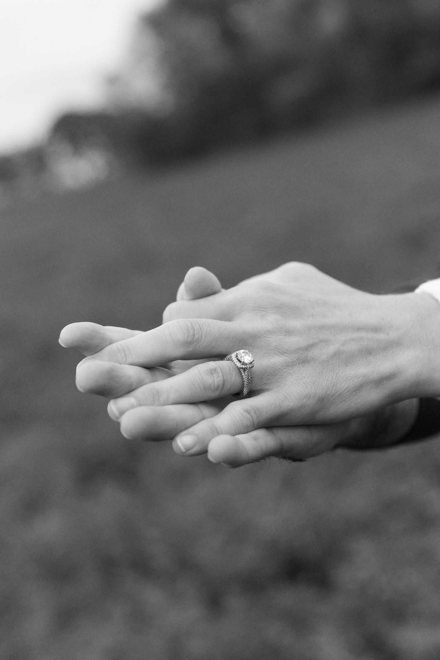 Black and white photo of two hands clasped together, one wearing an elegant engagement ring, outdoors.