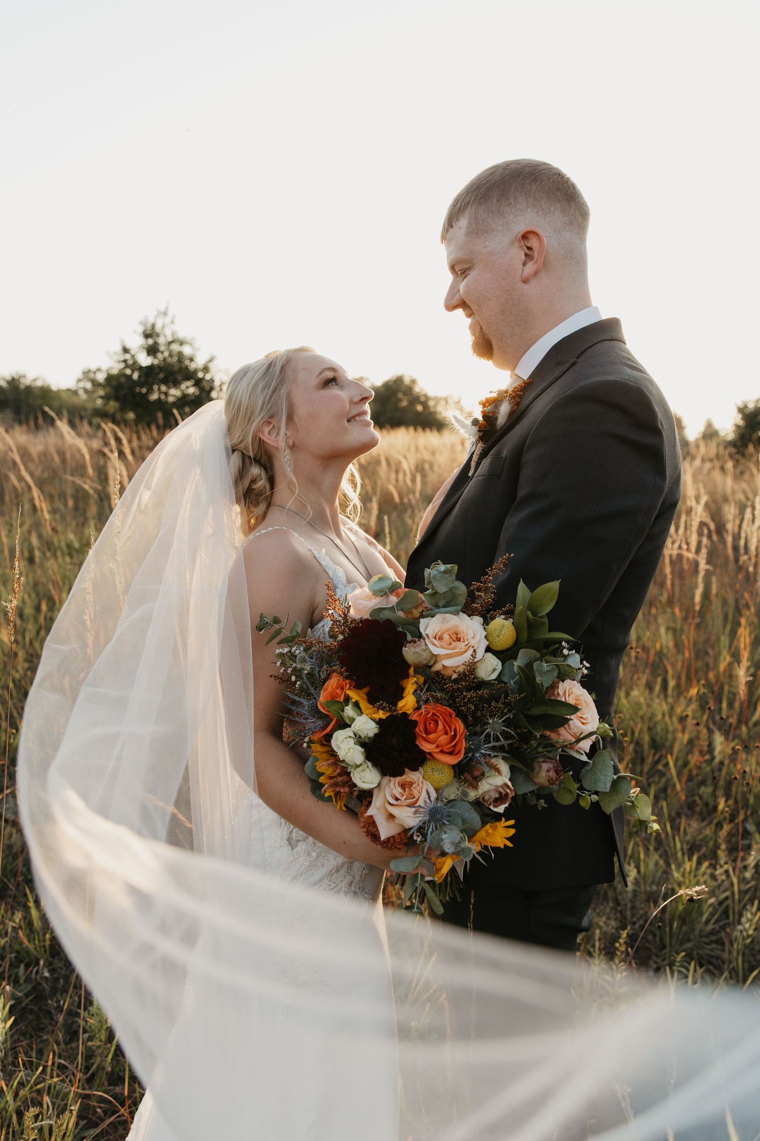 Bride and groom share a romantic moment in a golden field, her veil flowing as she holds a vibrant fall bouquet.