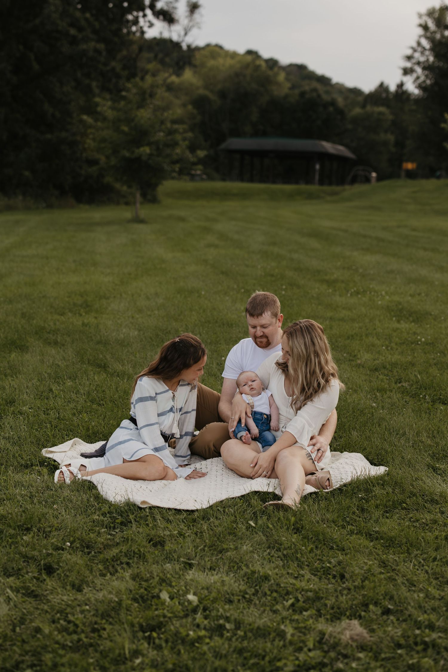 Family of four sitting on a blanket in a lush green park, parents lovingly attending to their baby girl.