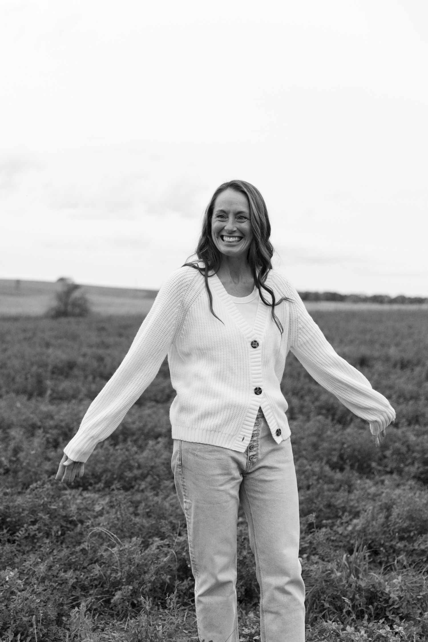 Black and white photo of a smiling woman in a knit cardigan and jeans standing in an open field.