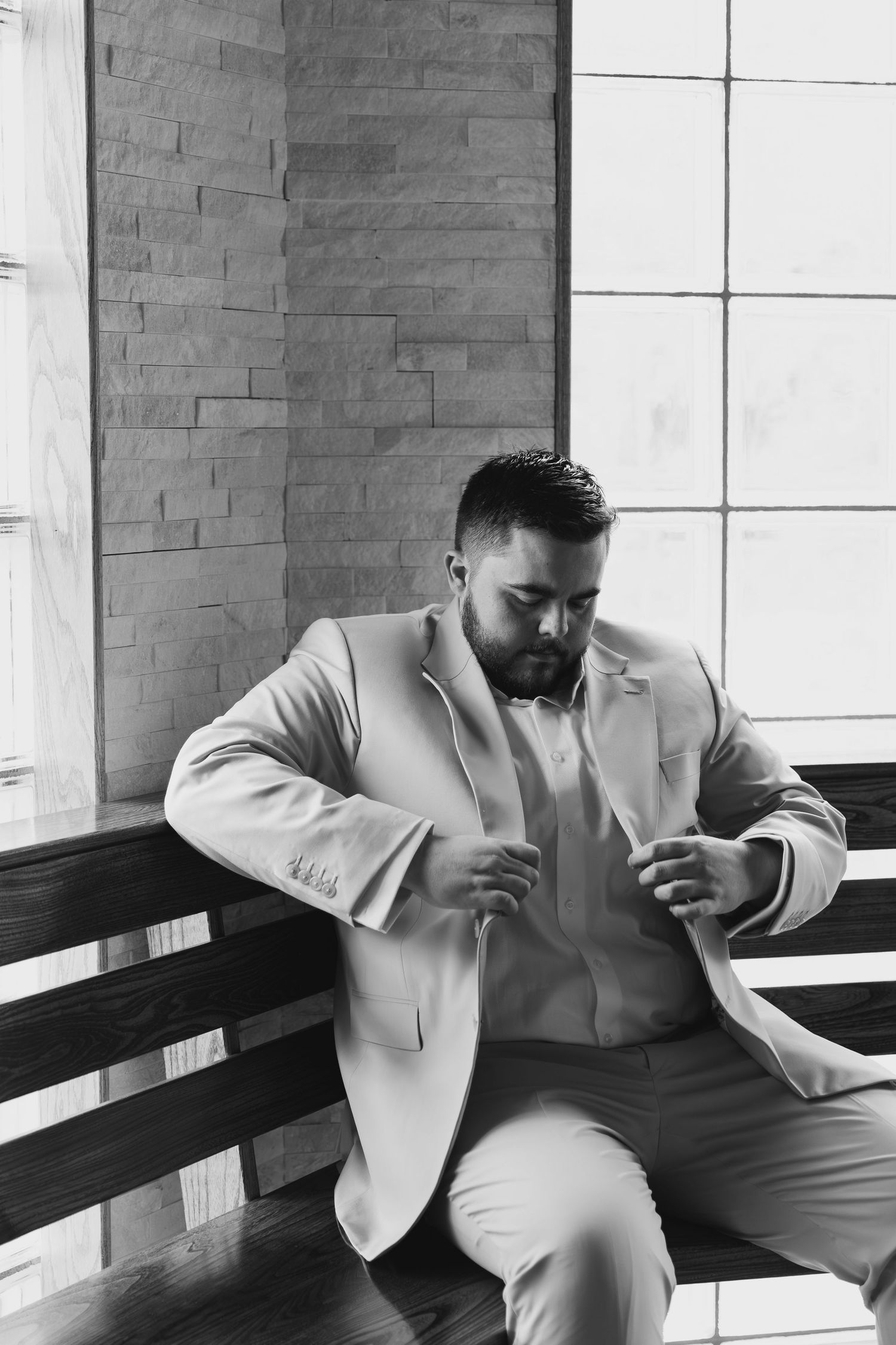 Black and white photo of a well-dressed man in a suit sitting on a bench, adjusting his jacket indoors.