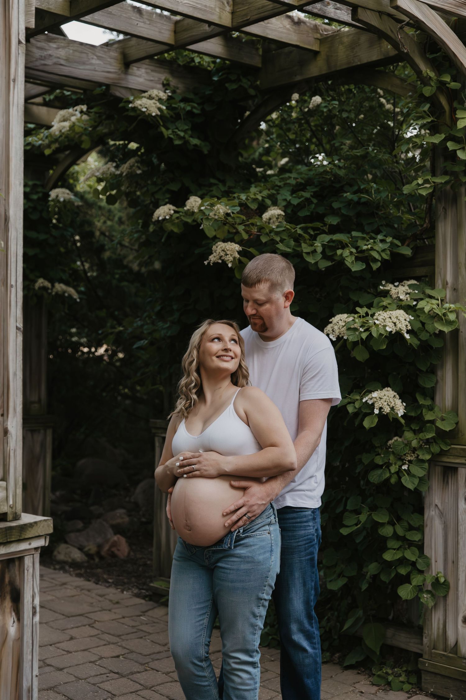 Pregnant couple embracing under wooden pergola surrounded by lush greenery and white hydrangeas during outdoor maternity photo session.