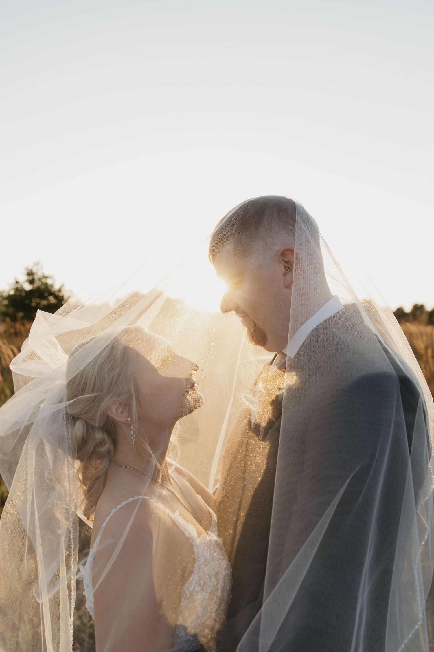 Bride and groom share a romantic moment under a flowing veil at golden hour in an outdoor field setting.