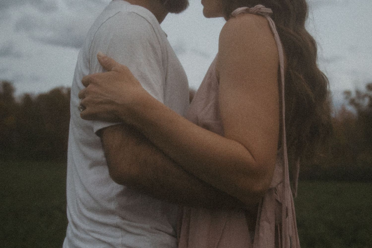 Romantic couple embracing outdoors in a field, woman in pink dress with long hair, moody cloudy sky background.
