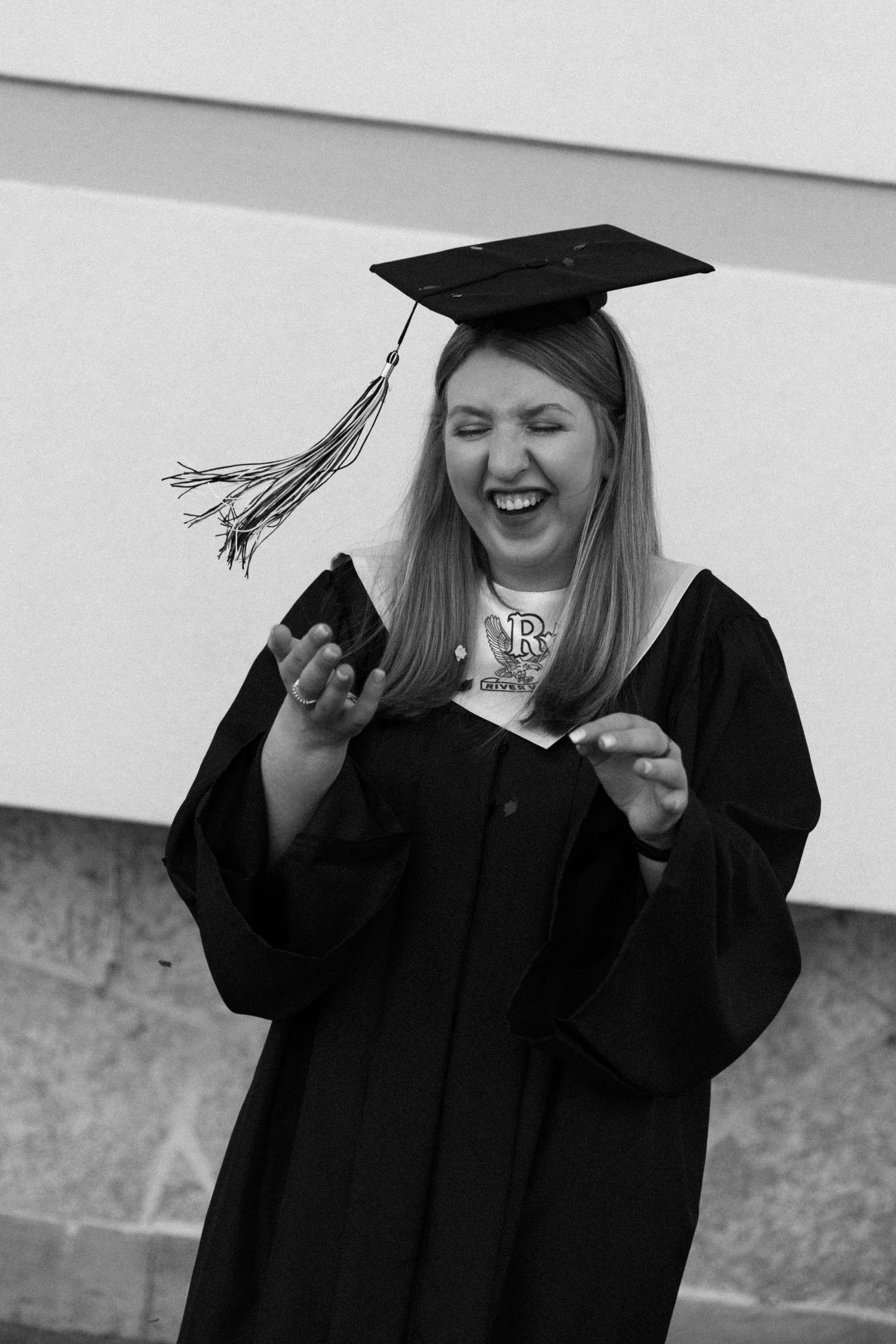 A joyful graduate in cap and gown laughs while holding a phone, celebrating her achievement in black and white.