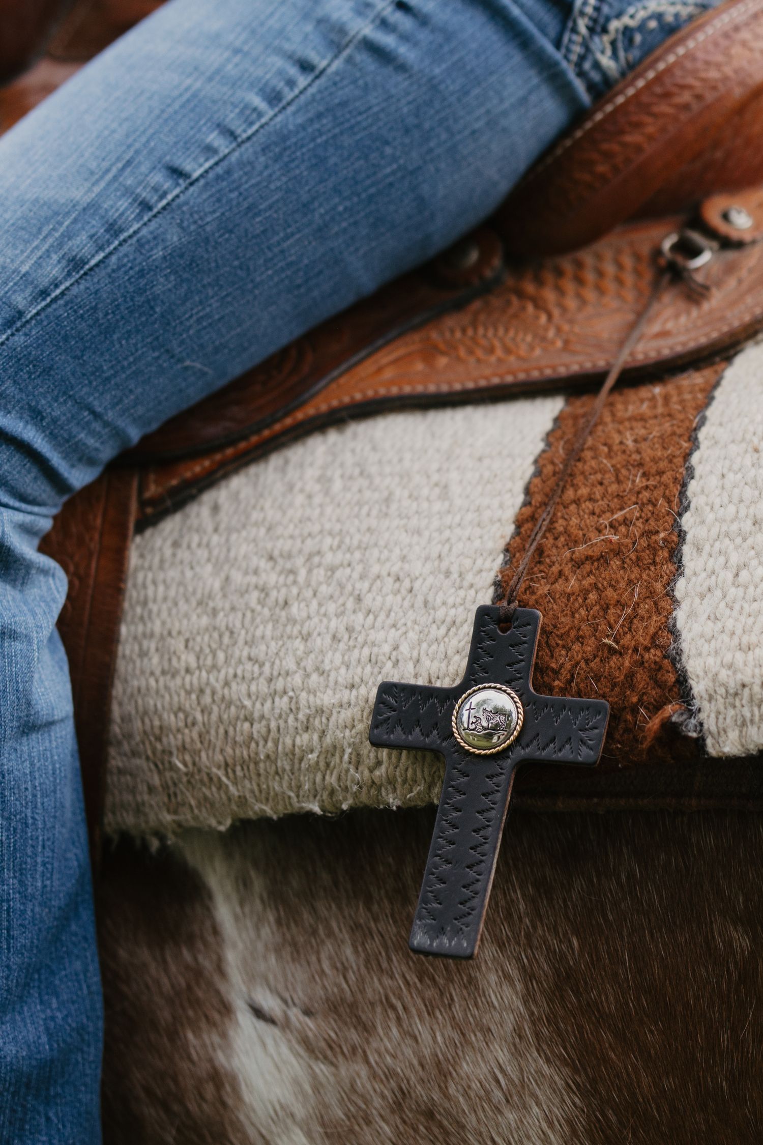 Dark leather cross pendant with silver concho detail resting on a Western saddle beside denim jeans.