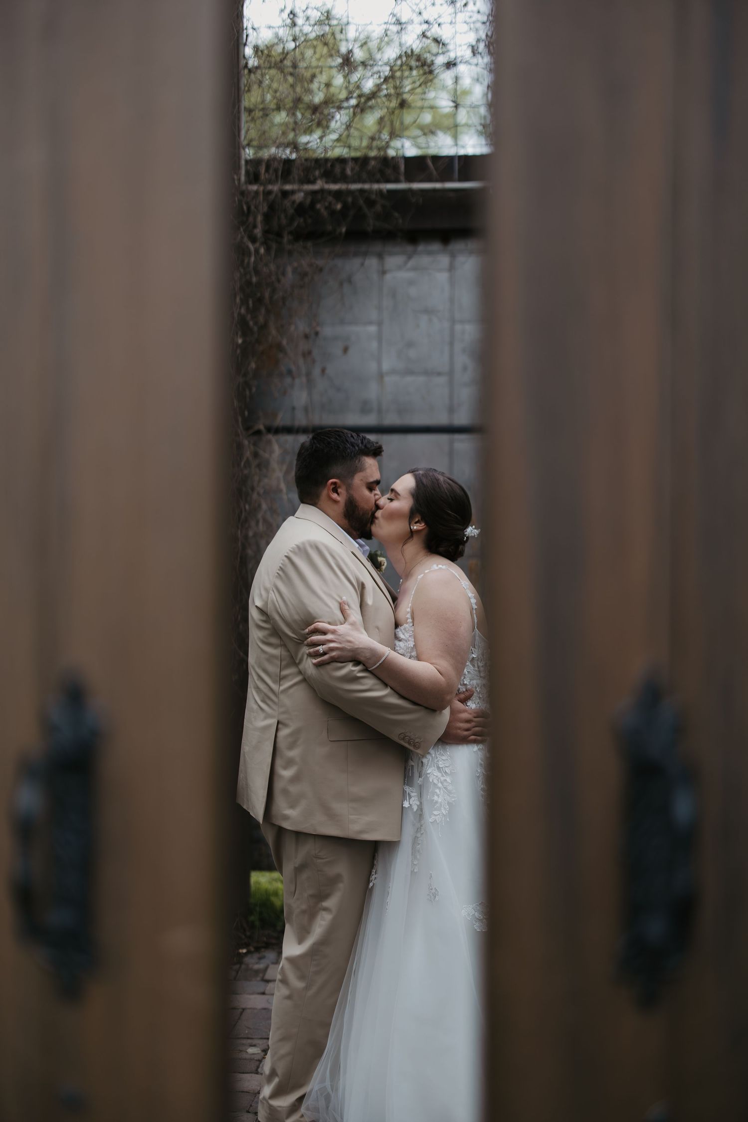 A bride and groom share a romantic kiss framed by wooden doors during their wedding day outdoor portrait.