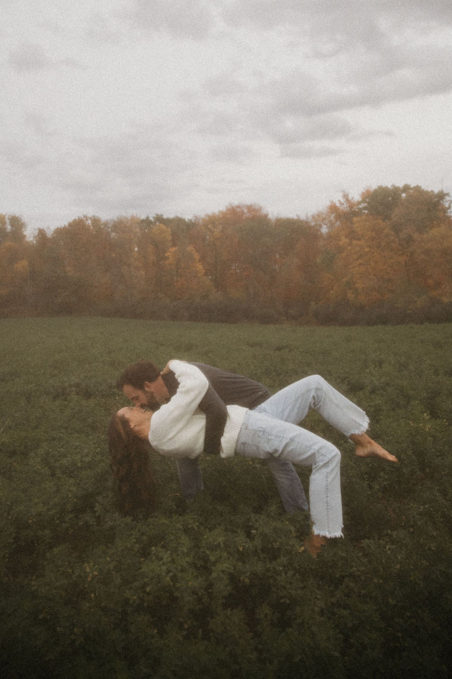 Couple sharing a romantic dip kiss in a green field with colorful autumn trees in the background.