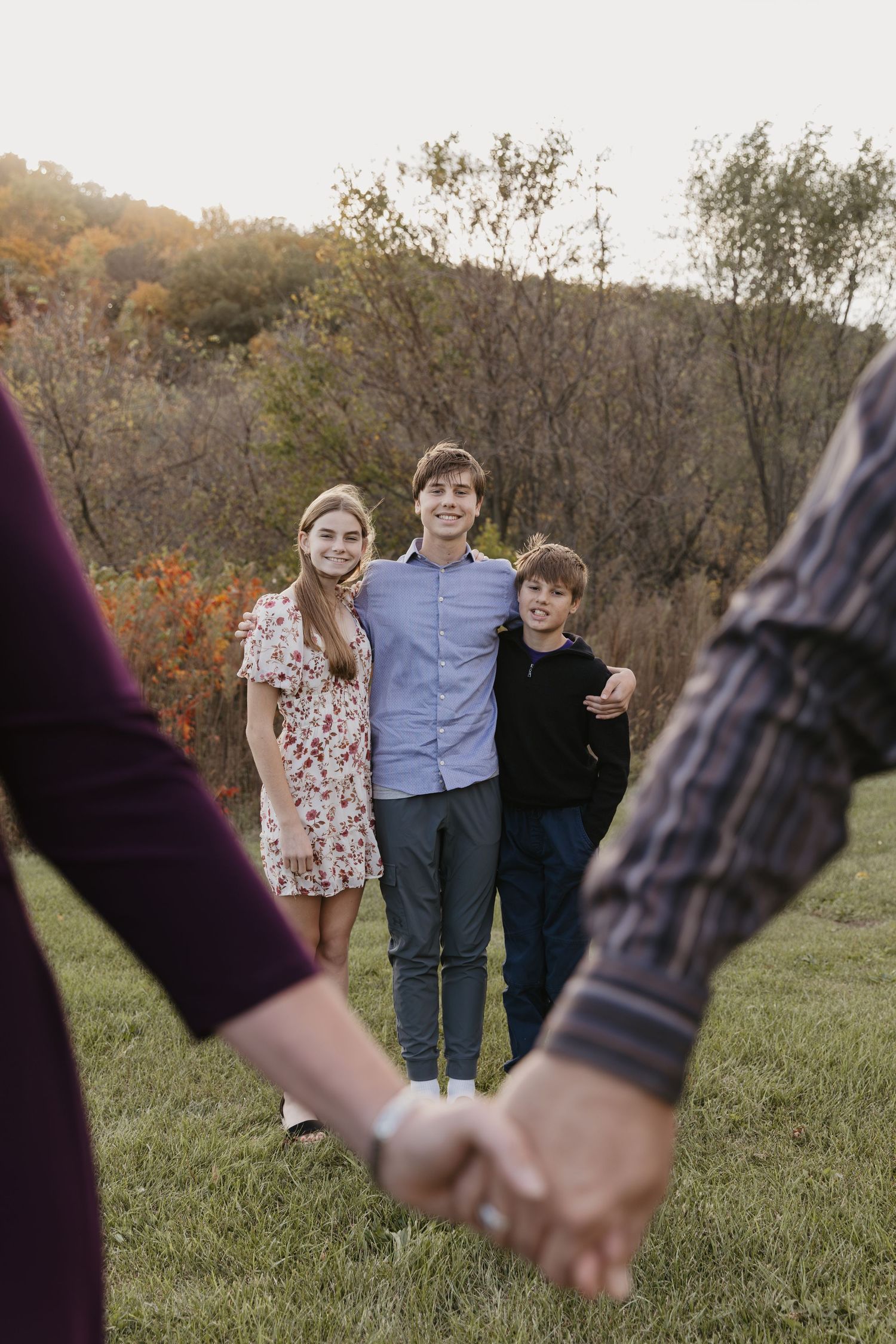 Three smiling siblings stand together on a grassy field with autumn trees, while two adults hold hands in the foreground.