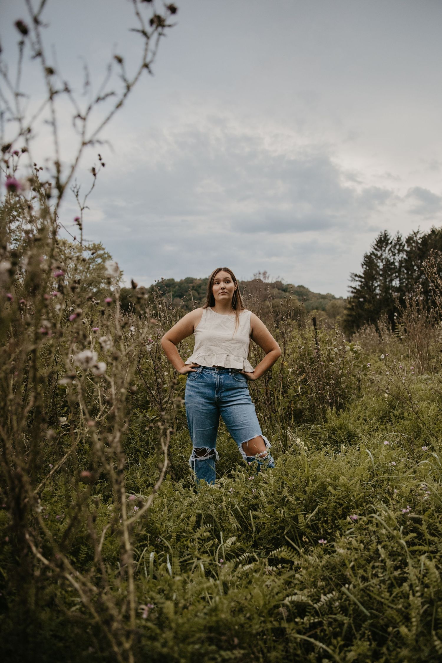 Young woman in ripped jeans and white top stands confidently in a wildflower meadow with cloudy skies.