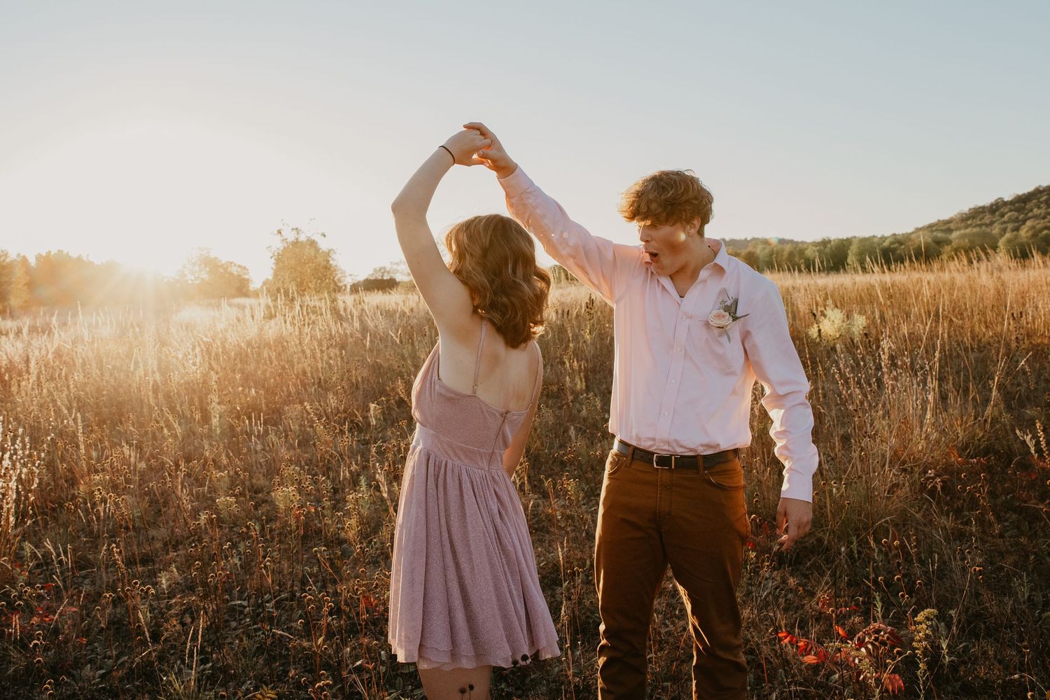 Couple dancing together in a golden sunlit field, man twirling woman in pink dress during romantic outdoor photo session.