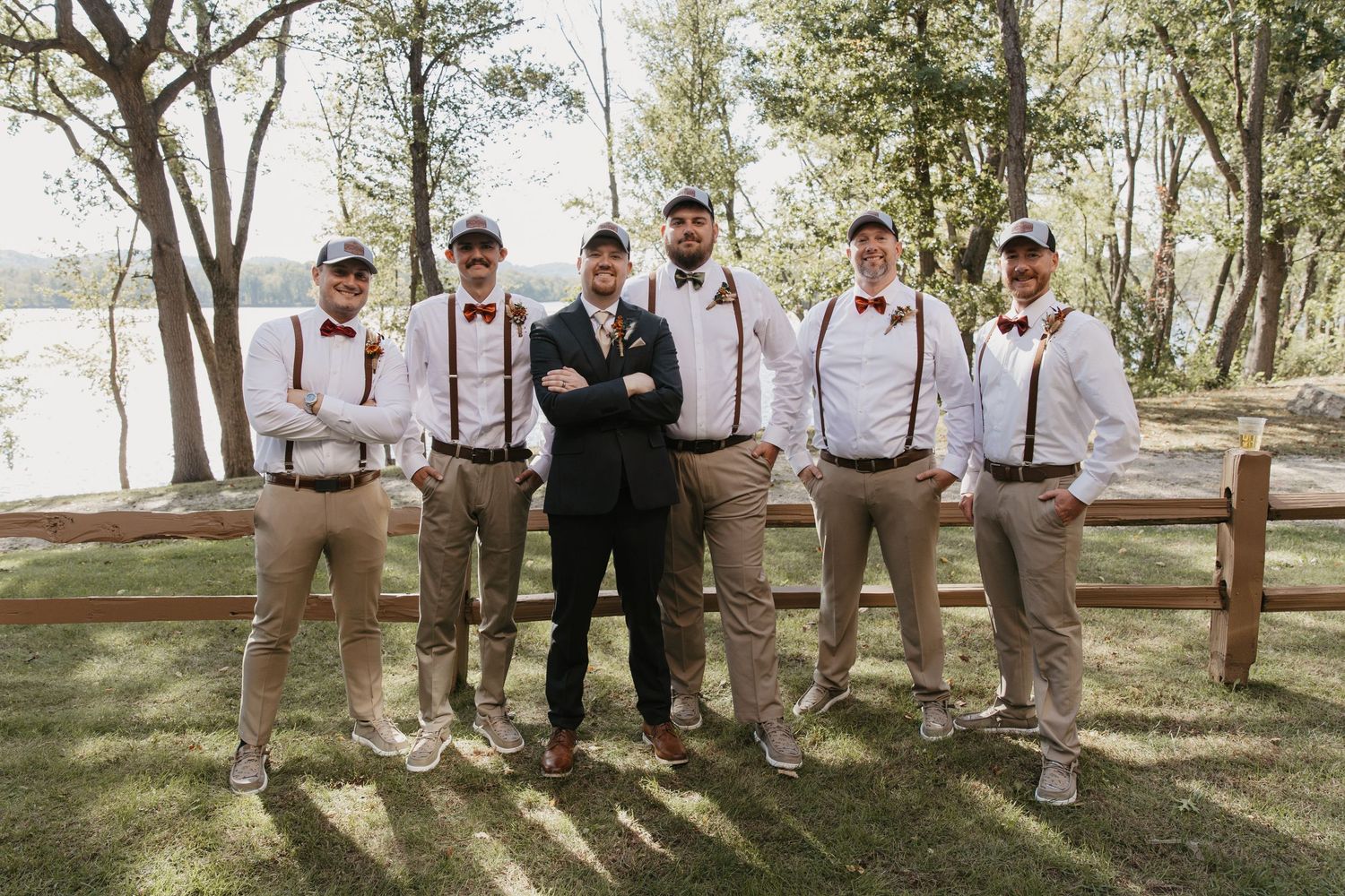 Groom and groomsmen in white shirts, suspenders, bow ties, and caps posing outdoors by a wooden fence.