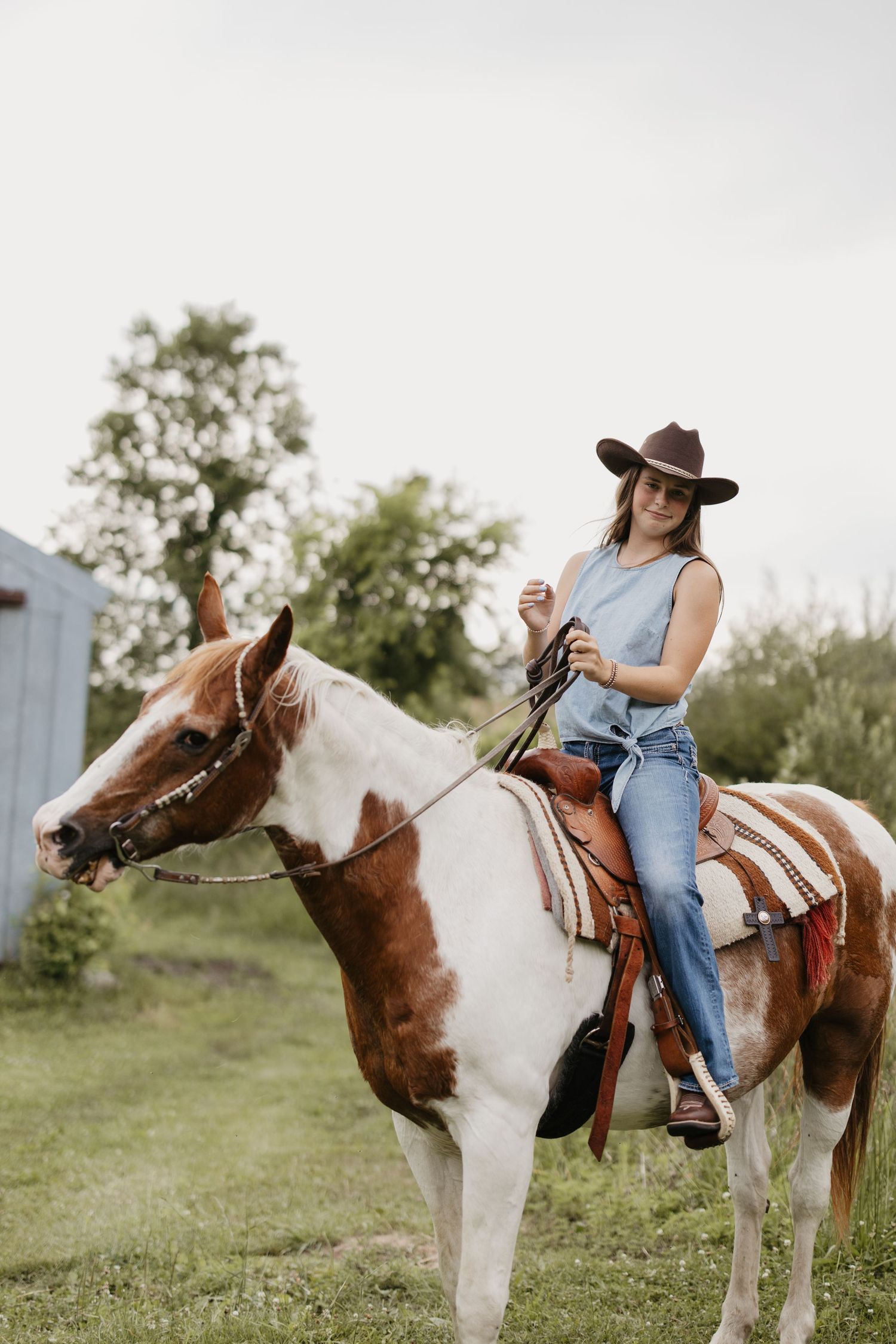 Woman in a brown cowboy hat and denim outfit riding a brown and white paint horse across a green grassy field.