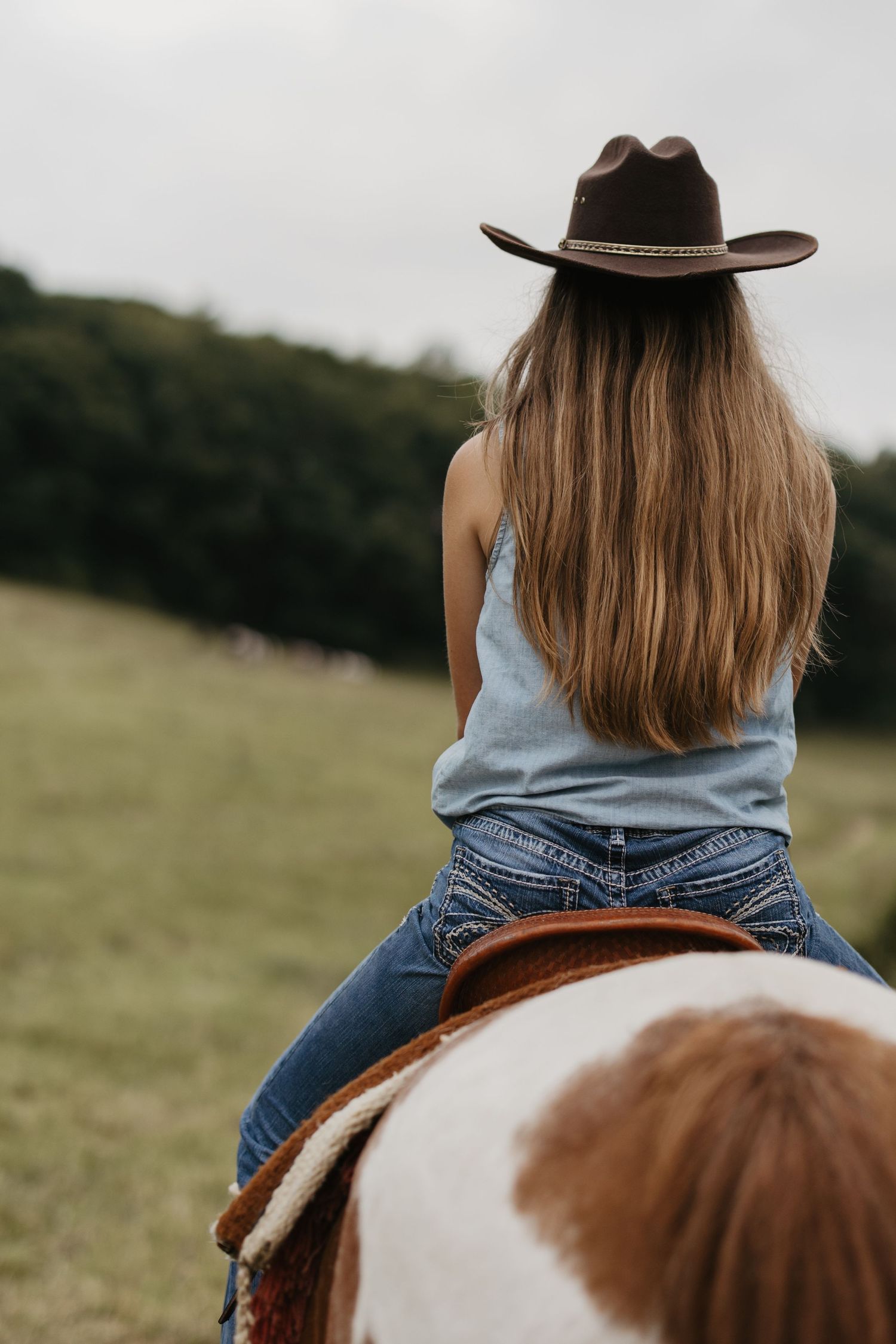 Woman in cowboy hat and jeans riding a paint horse through a green field, viewed from behind.