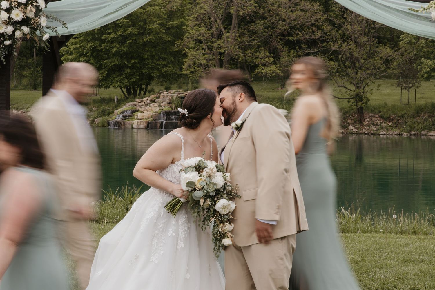 Bride and groom share a kiss outdoors near a pond, surrounded by wedding party in sage green and tan attire.