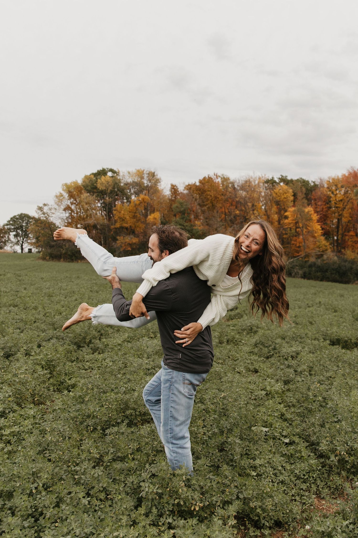 A joyful couple plays in a green field with autumn trees, man lifting laughing woman in a fun outdoor fall photo.