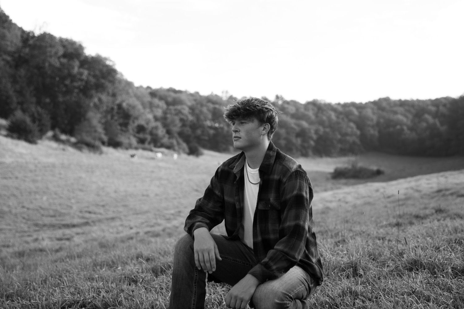 Young man in plaid shirt sitting on grass in black and white rural landscape photo.