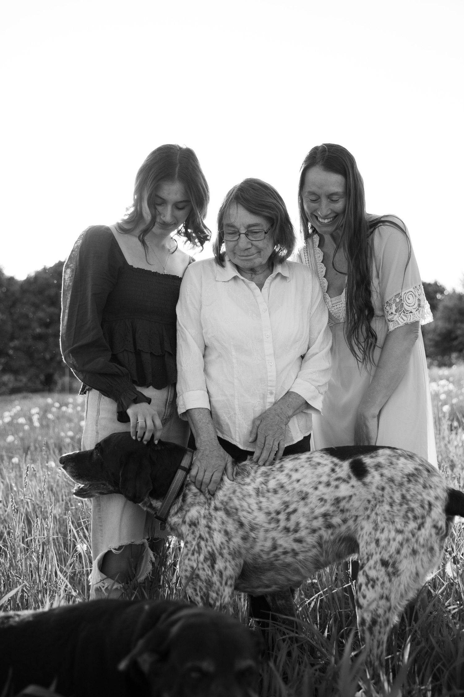 Black and white photo of two young women and an elderly woman smiling while petting dogs in a field.