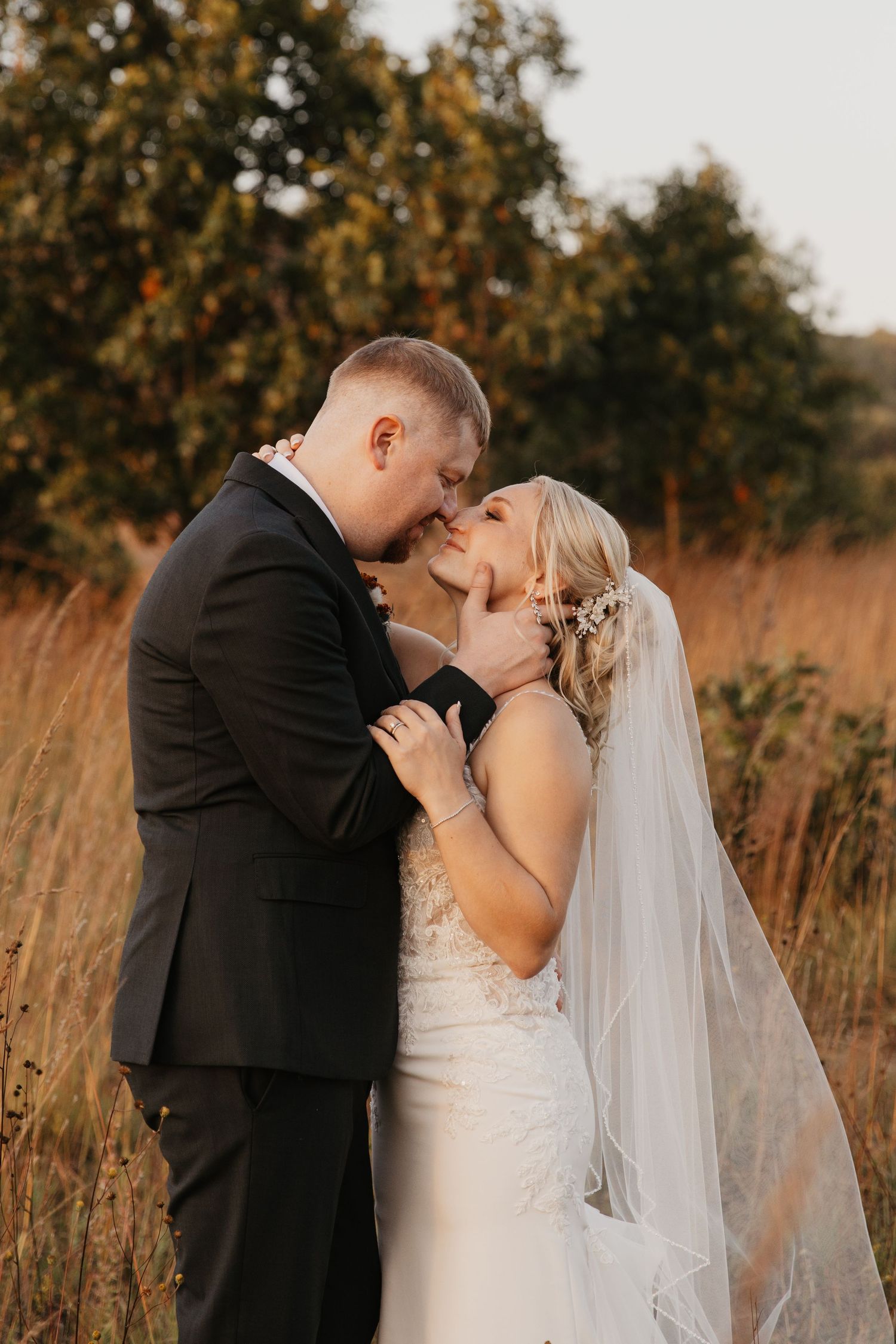 Bride and groom share a romantic kiss in a golden field at sunset, bride wearing lace gown and veil.