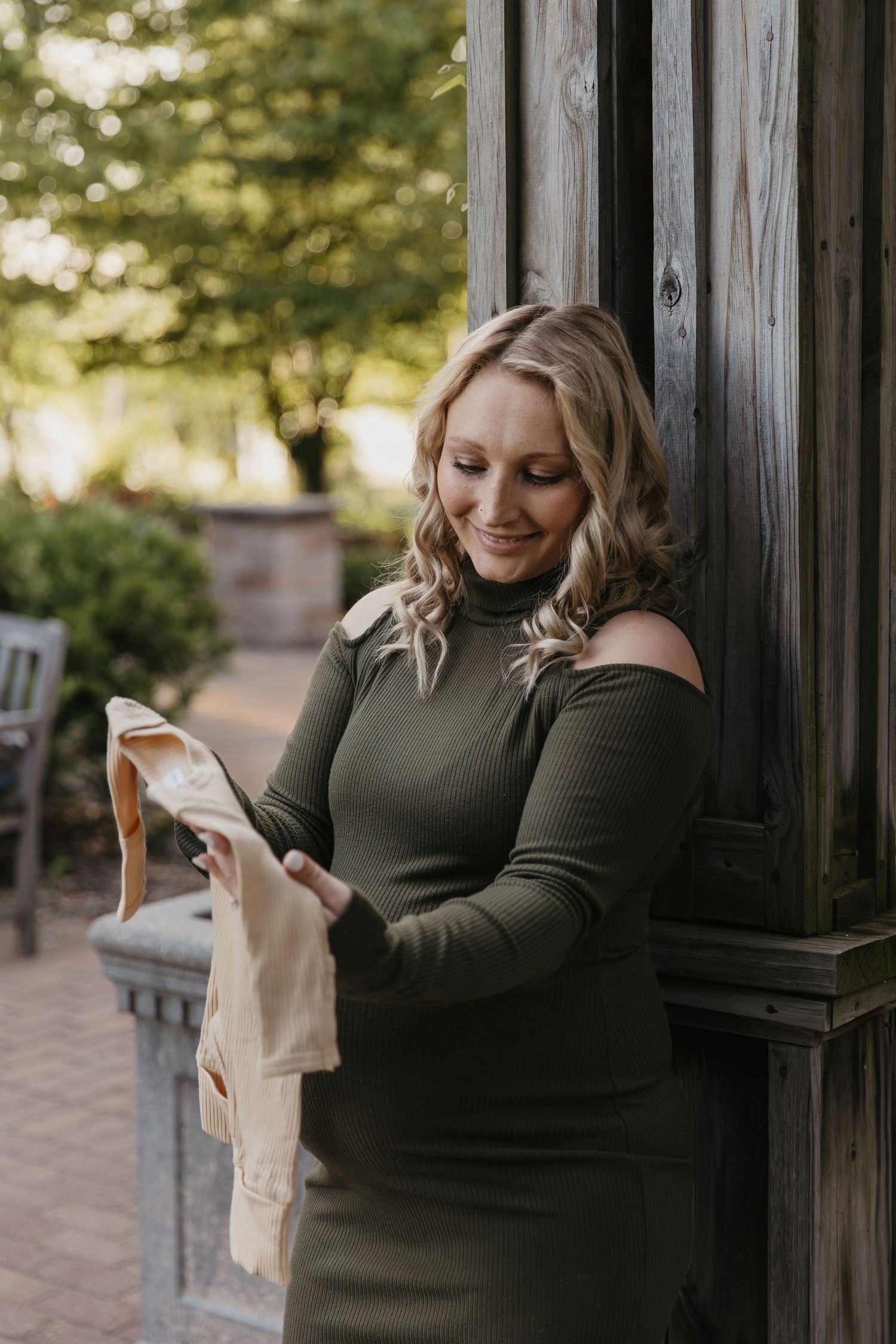 Smiling blonde woman in olive green off-shoulder dress holding baby clothes outdoors near wooden pillar.