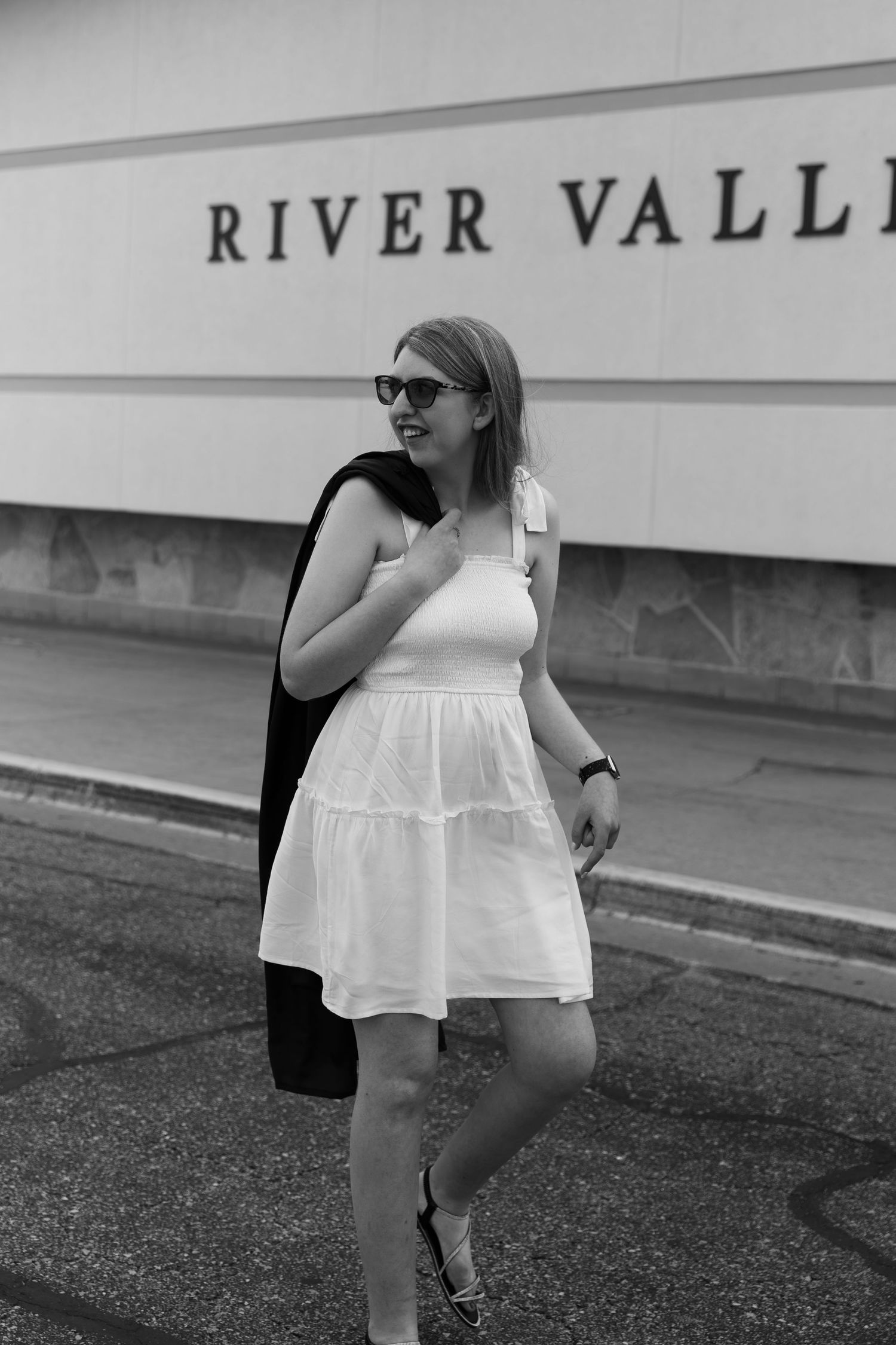A young woman in a white sundress and sunglasses smiles while walking in front of a River Valley sign.