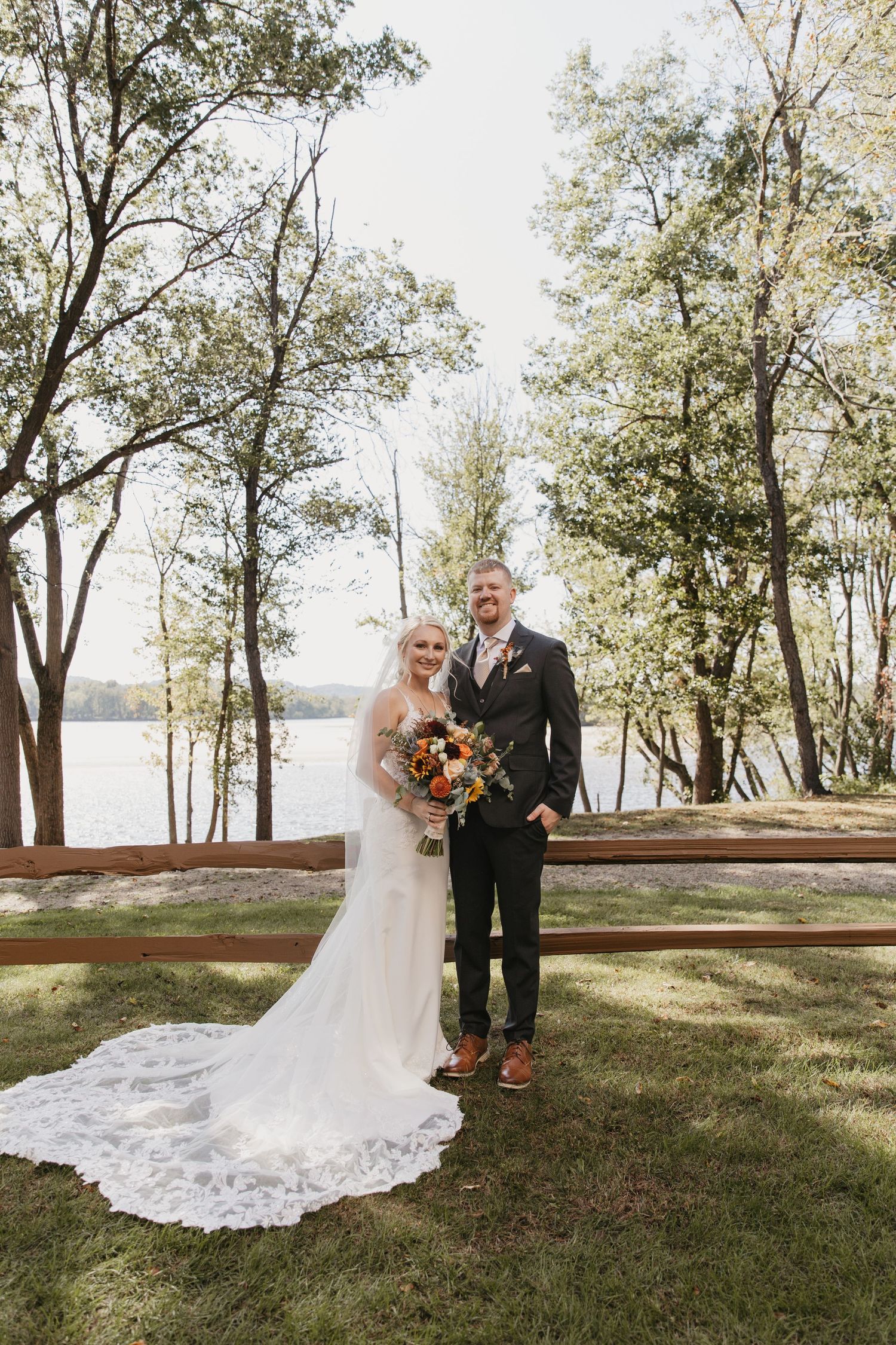 Bride and groom pose together outdoors near a lakeside wooden fence, surrounded by trees, bride holding colorful bouquet.