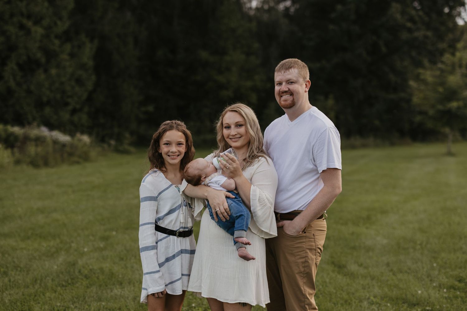 Family of four posing outdoors on green grass, parents holding baby boy with young girl smiling beside them.