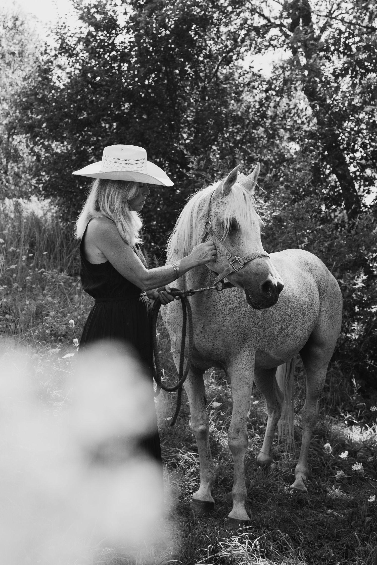 Black and white photo of a blonde woman in a sun hat and dress standing beside a white horse outdoors.