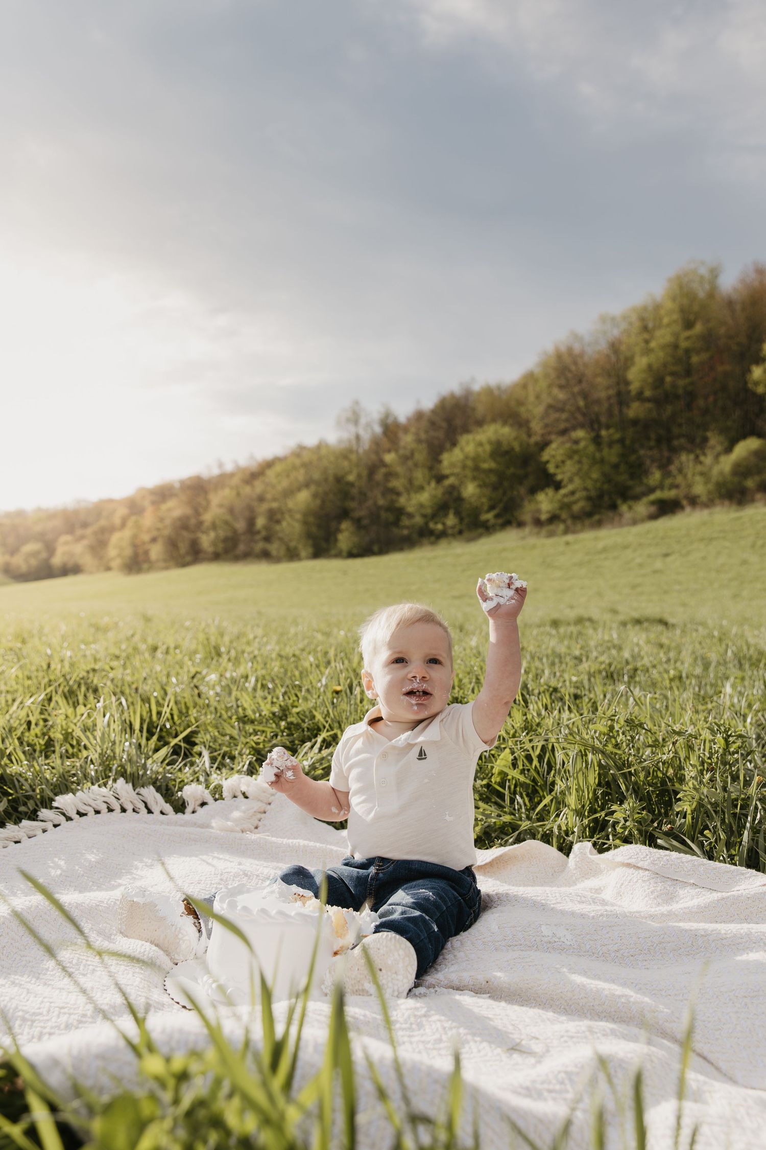 Happy baby sitting on white blanket in green field, raising arm joyfully on sunny day.