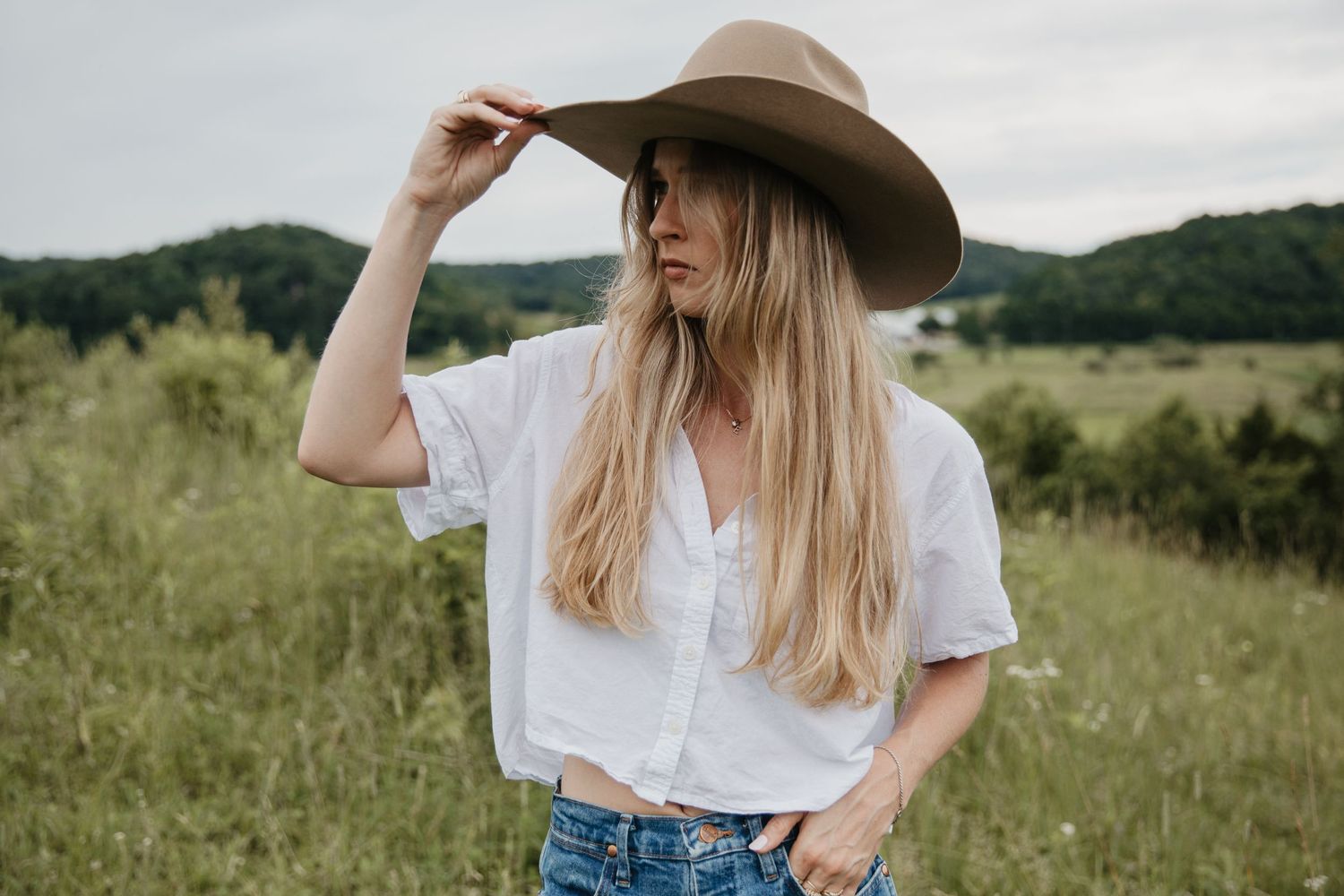 Person in casual attire and cowboy hat poses in a green summer field.