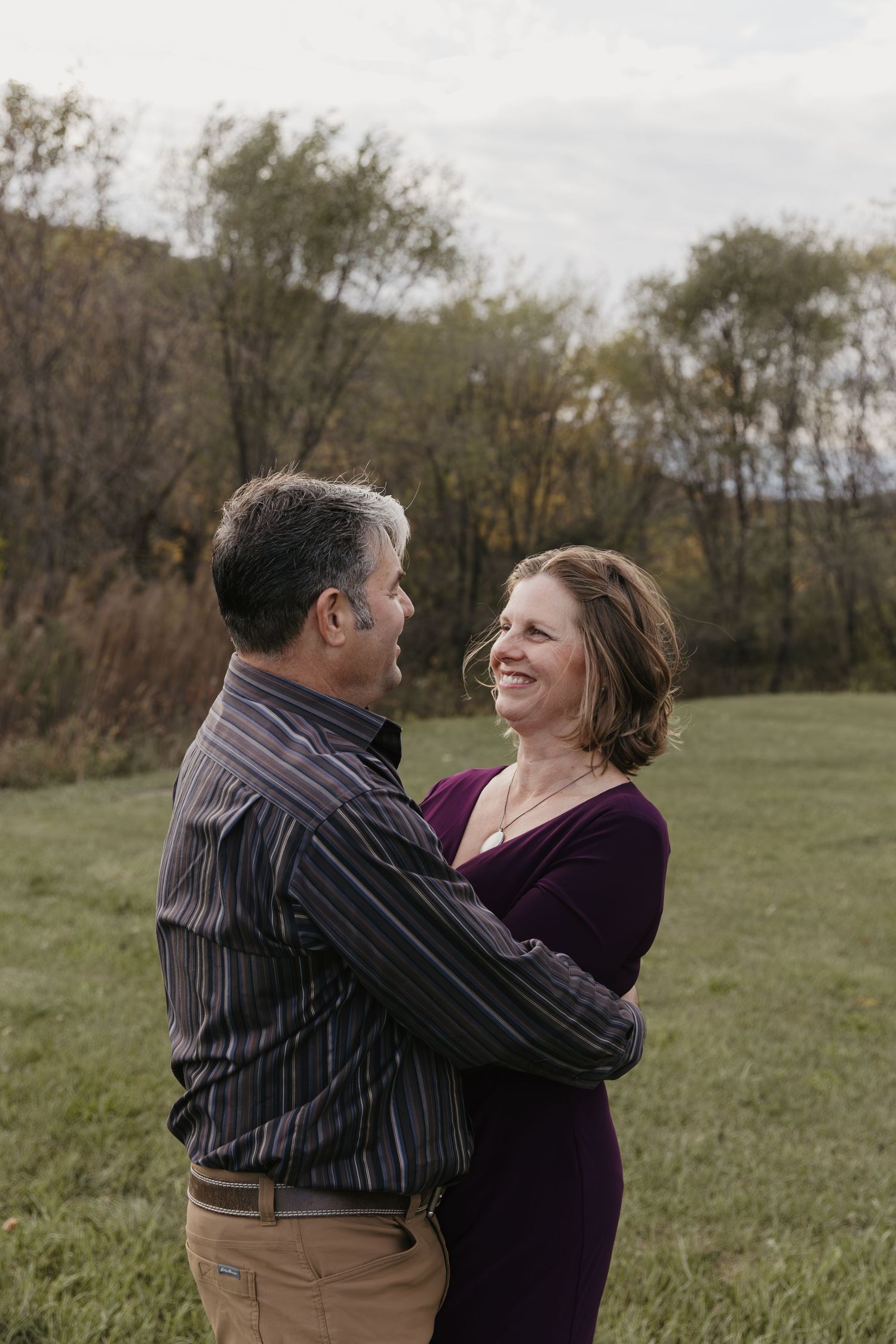 A couple embraces and smiles at each other in a grassy field with autumn trees in the background.
