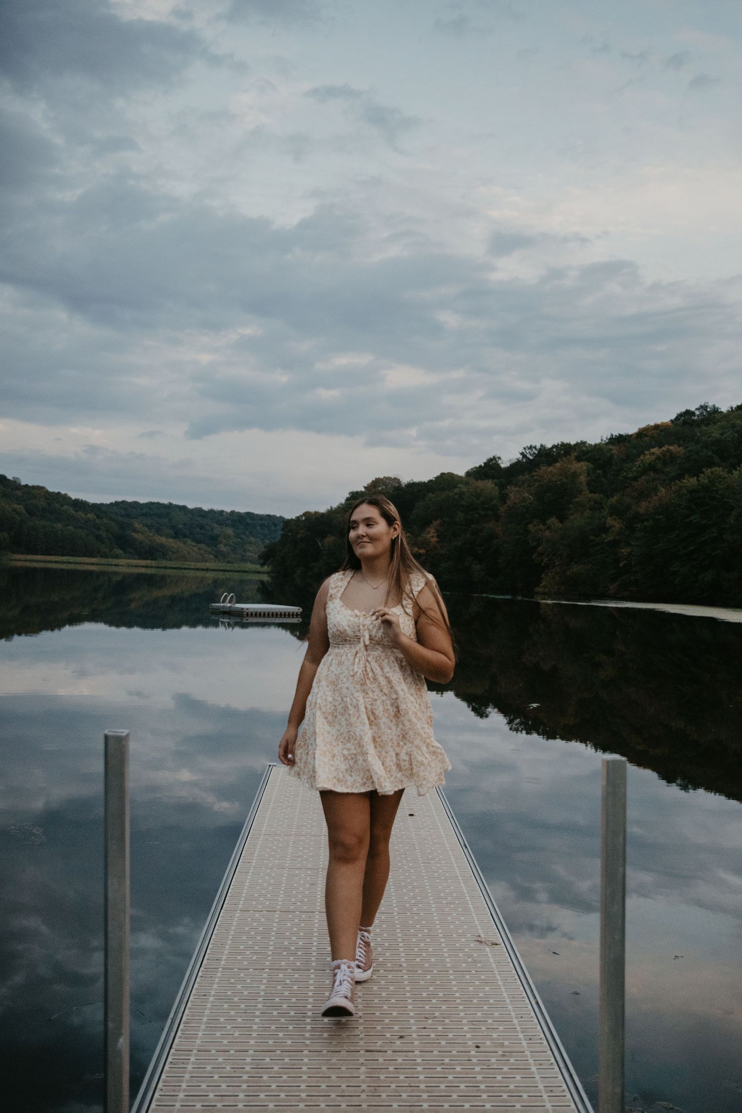 Young woman in floral dress walks on lake dock, trees reflecting in calm water under cloudy sky.