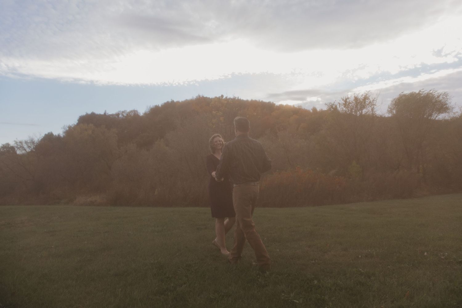Couple walking hand in hand across a green field at dusk with autumn trees in the background.