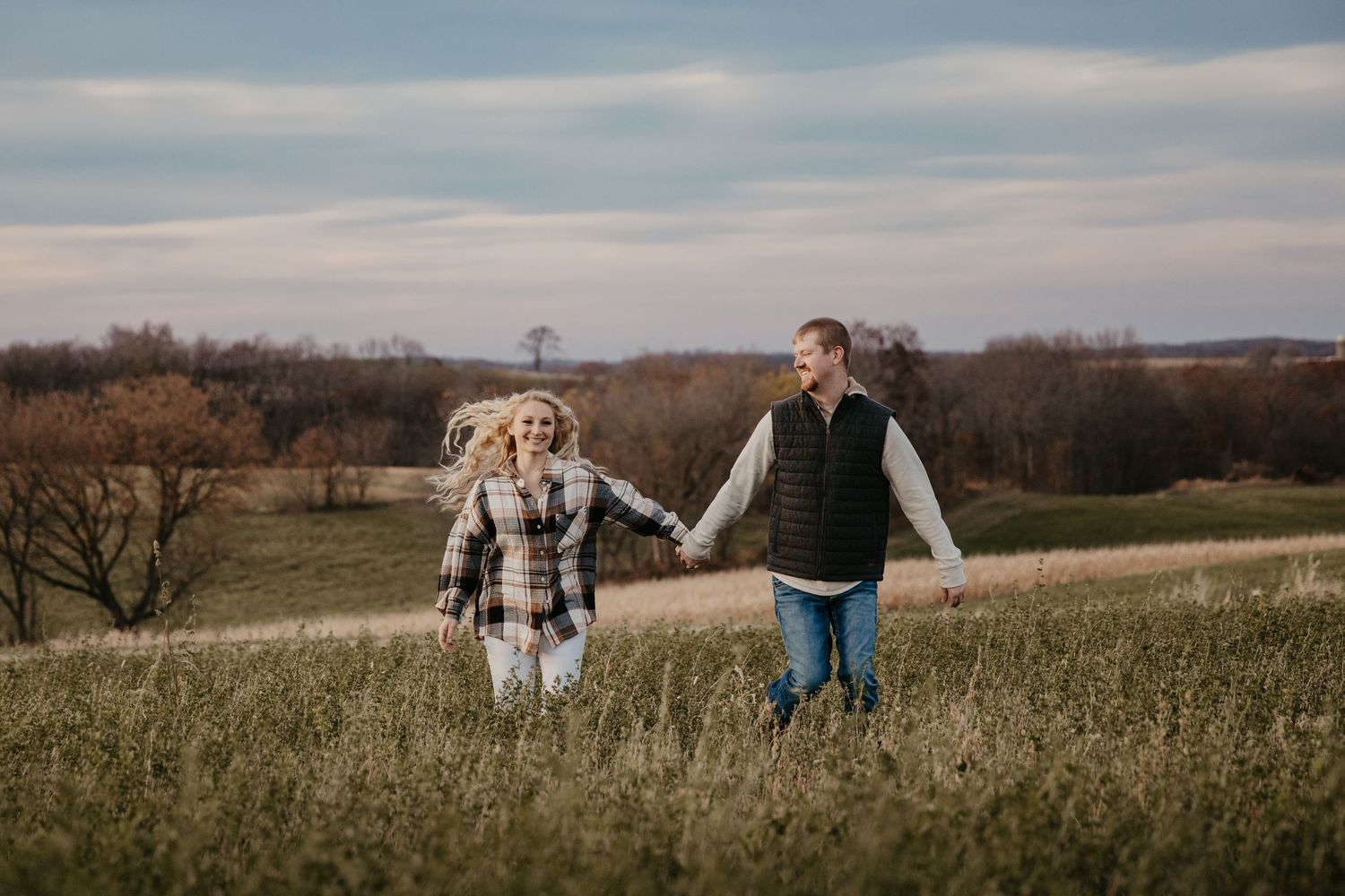 Couple holding hands walking through a green field with rolling hills and cloudy sky in a romantic outdoor engagement photo.