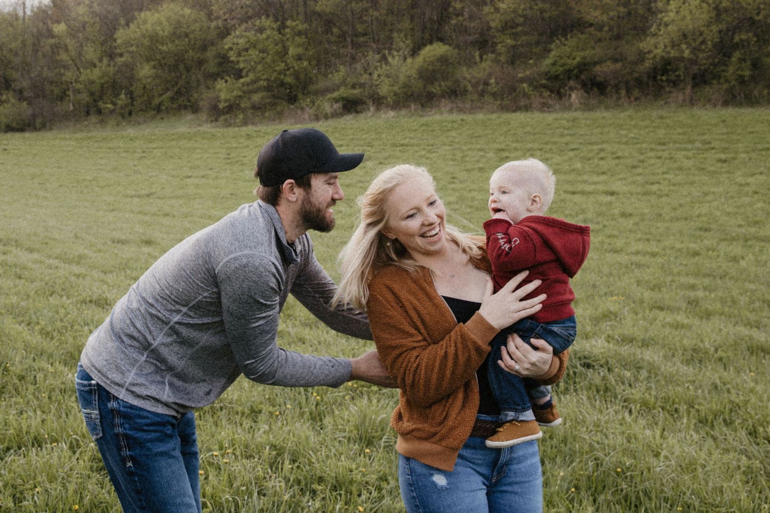 Happy family with parents and baby laughing together in a lush green field surrounded by trees.