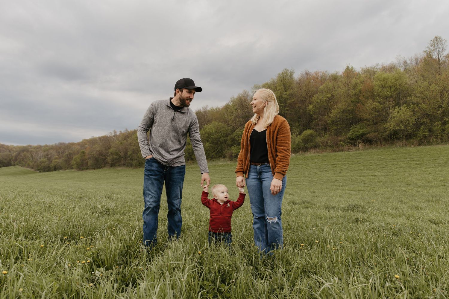 A young family with a toddler holds hands in a lush green field with autumn trees under a cloudy sky.