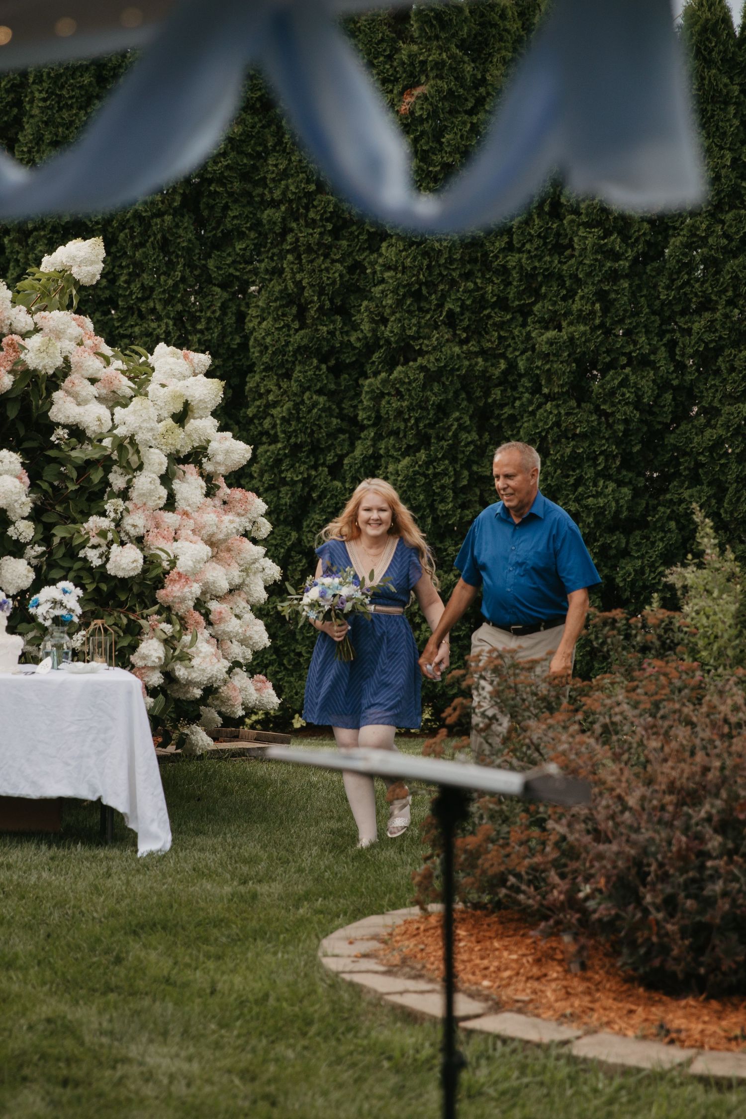 Couple in blue attire walks hand-in-hand through an outdoor garden wedding ceremony with white floral arrangements.