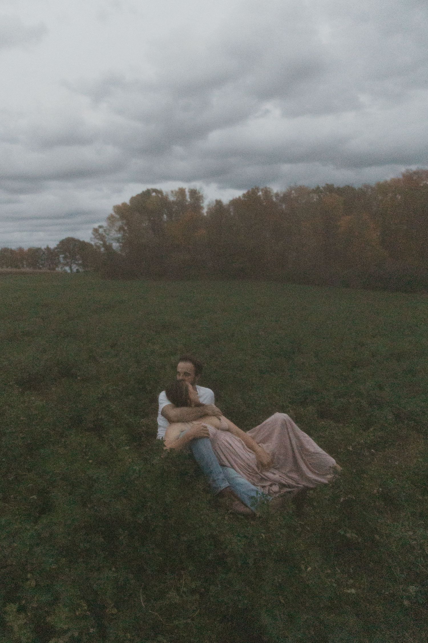 Couple sitting together in a lush green field wearing a pink dress and white shirt under cloudy autumn skies.