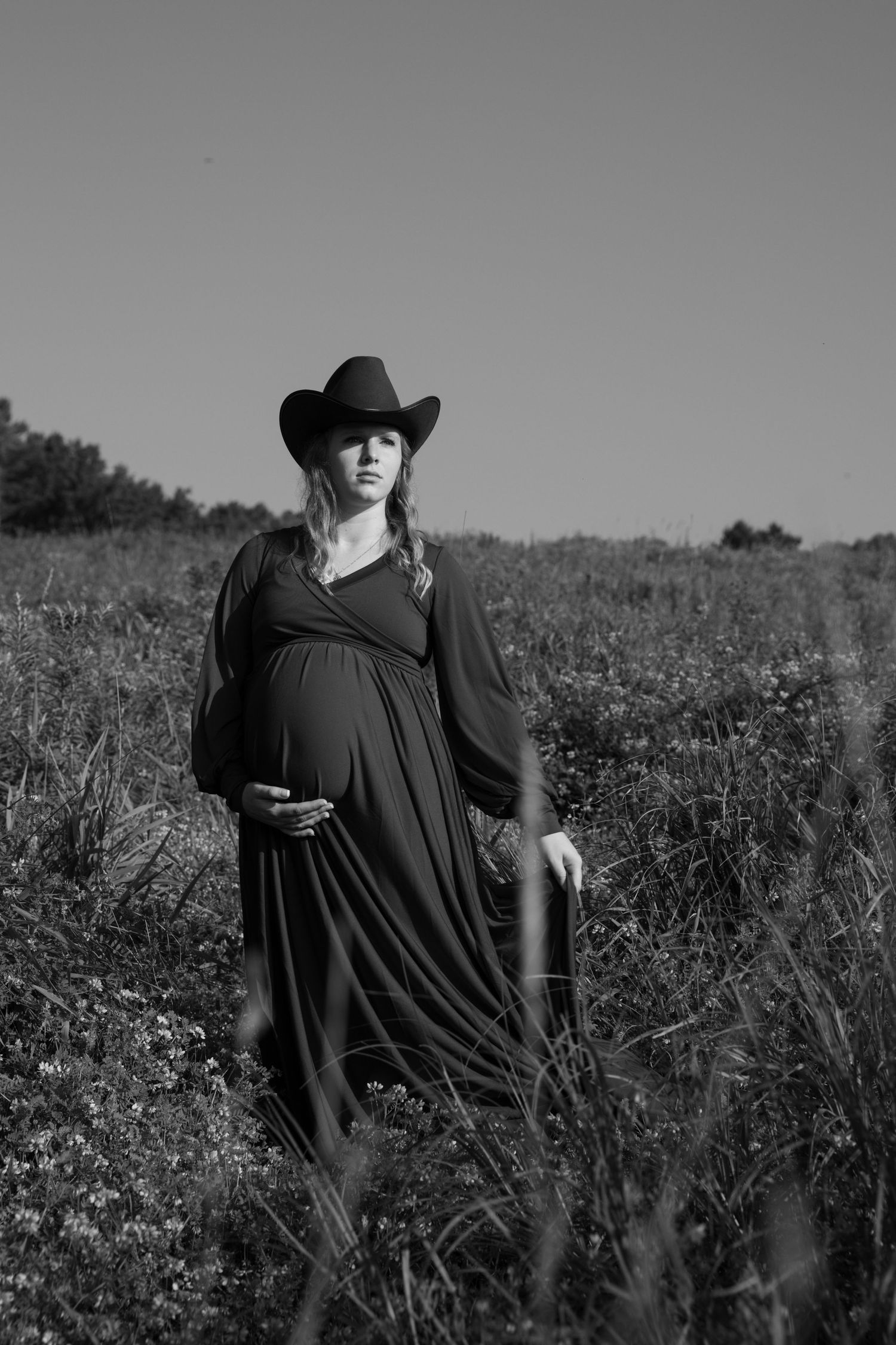 Black and white photo of a pregnant woman in a long dress and cowboy hat standing in a field.
