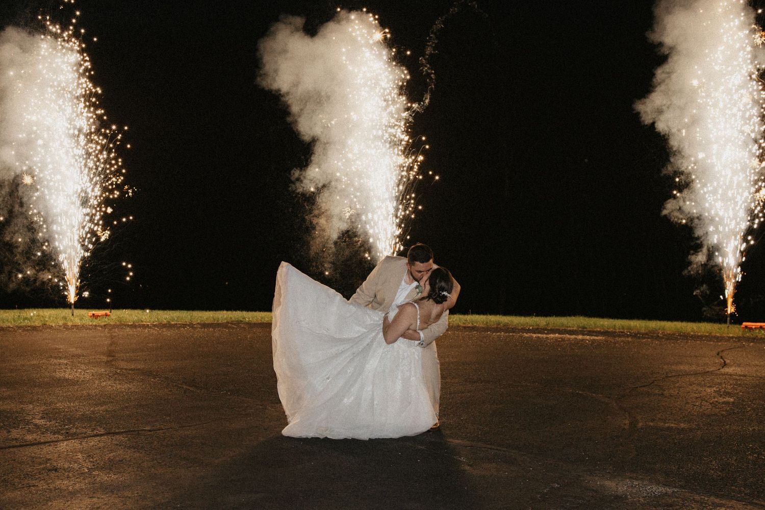 Bride and groom share a romantic dip kiss with sparkling fireworks fountains illuminating the night sky behind them.