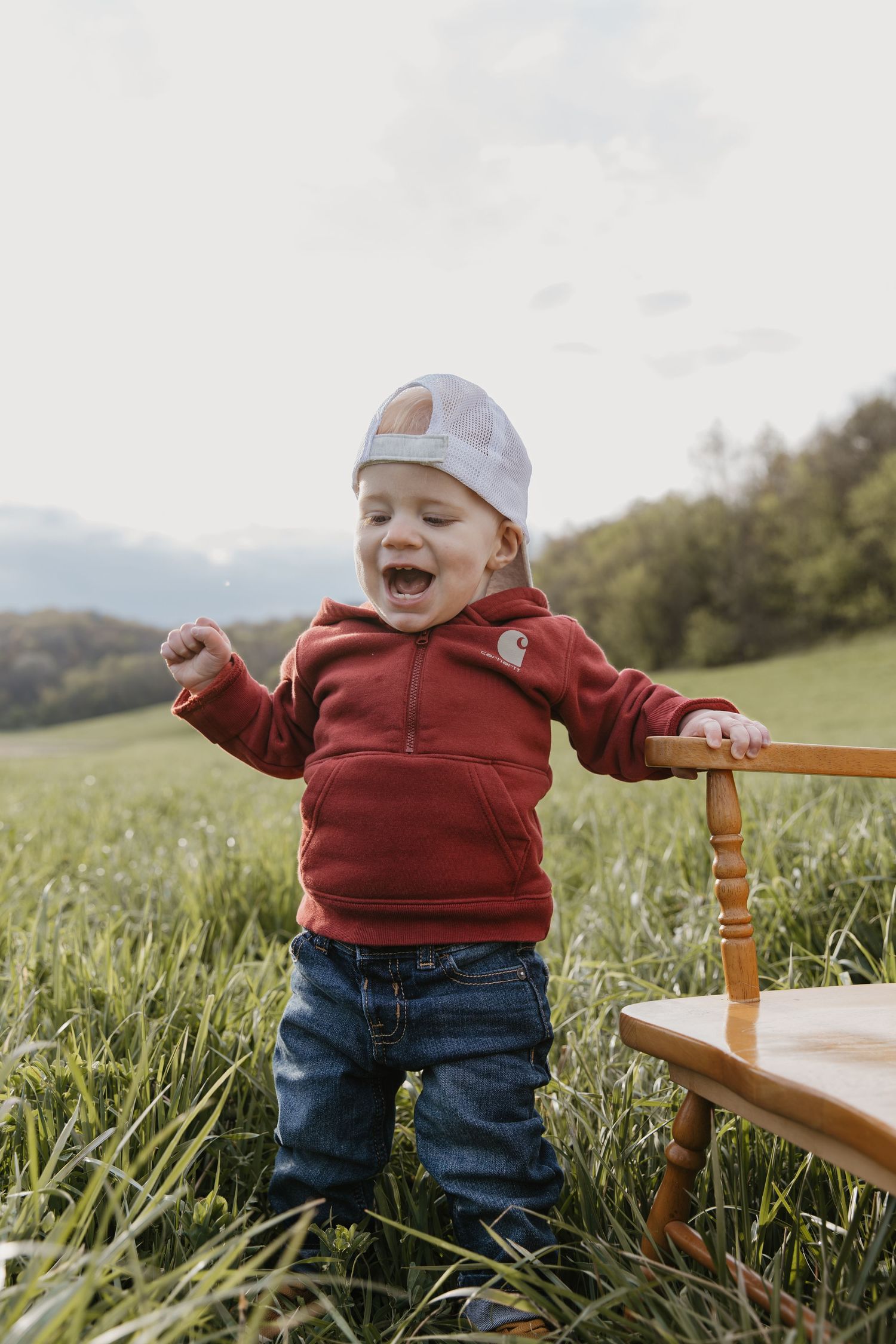 Joyful toddler in red Carhartt hoodie and jeans standing in green field holding wooden chair, wearing white trucker hat.