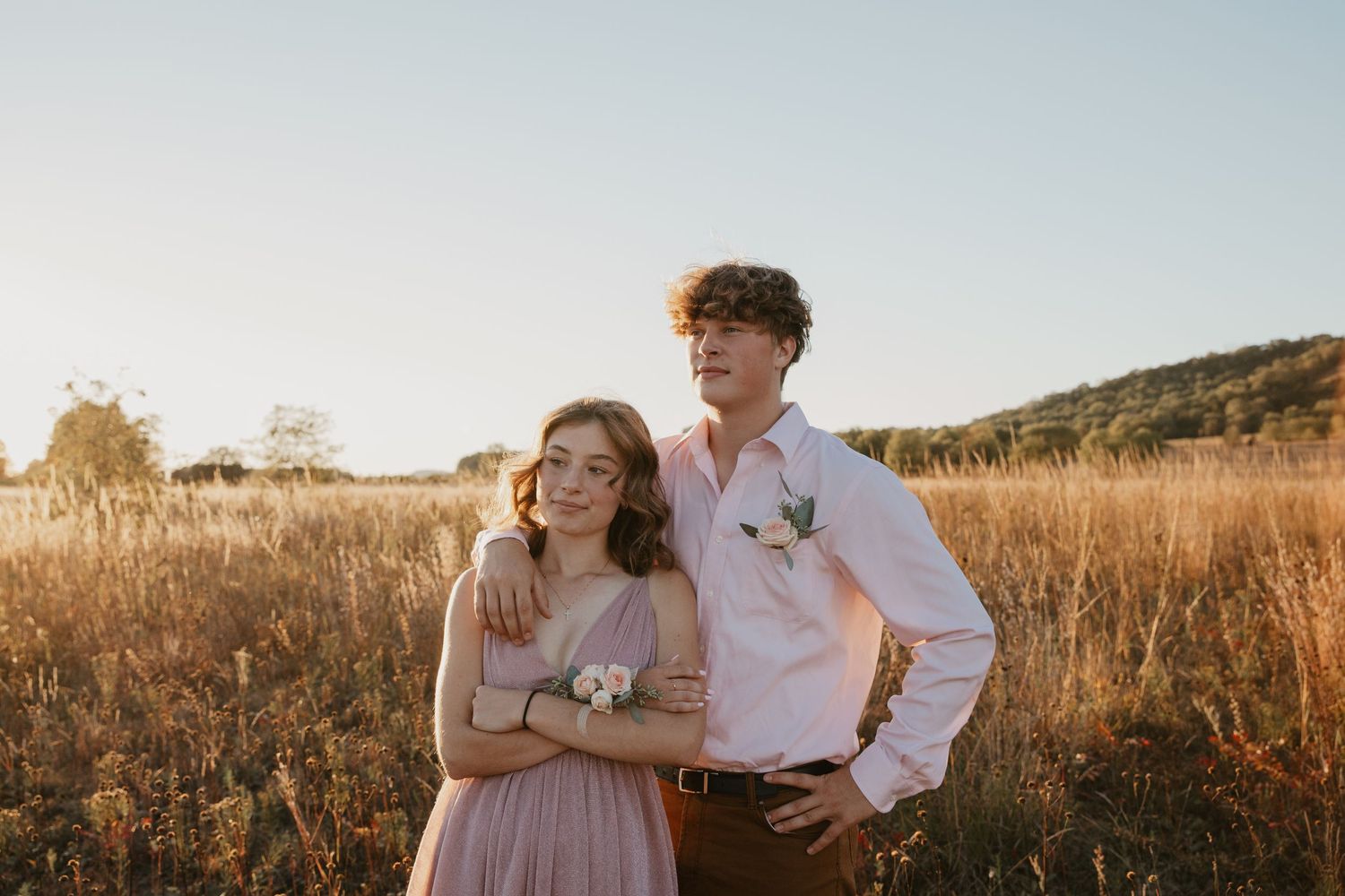 A young couple poses in a golden field at sunset, the girl in a pink dress and the boy in a white shirt with a boutonniere.