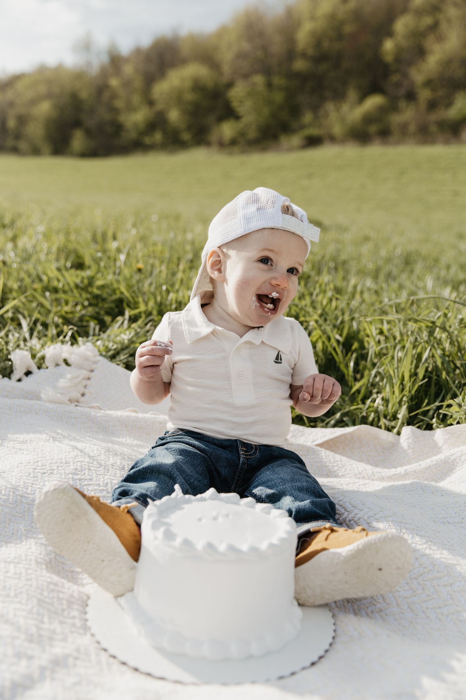 Happy toddler in a white cap and polo sits on a blanket in a grassy field, enjoying a small white smash cake.