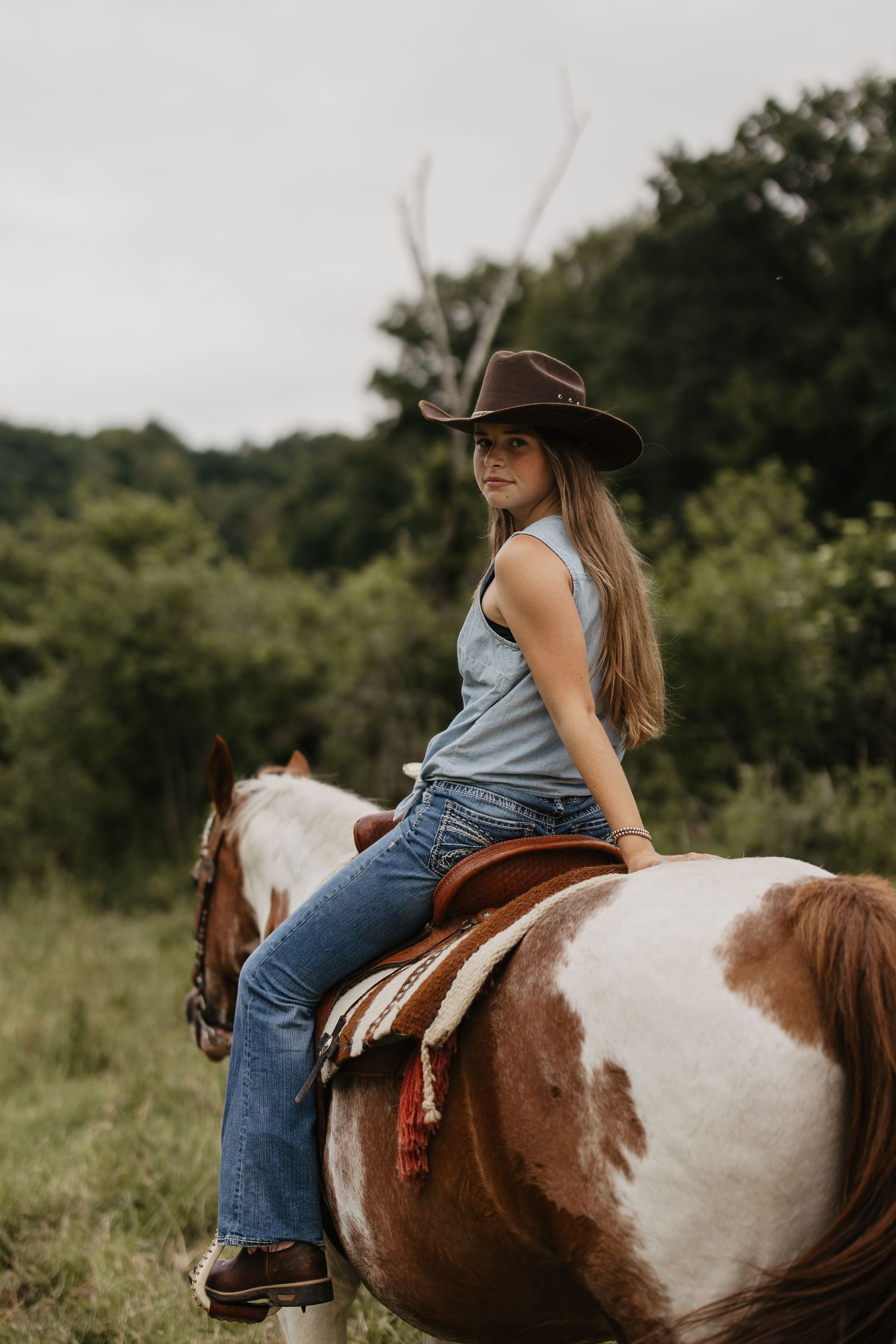 Young woman in a brown cowboy hat and denim jeans sits on a paint horse, looking back over her shoulder in a green countryside.